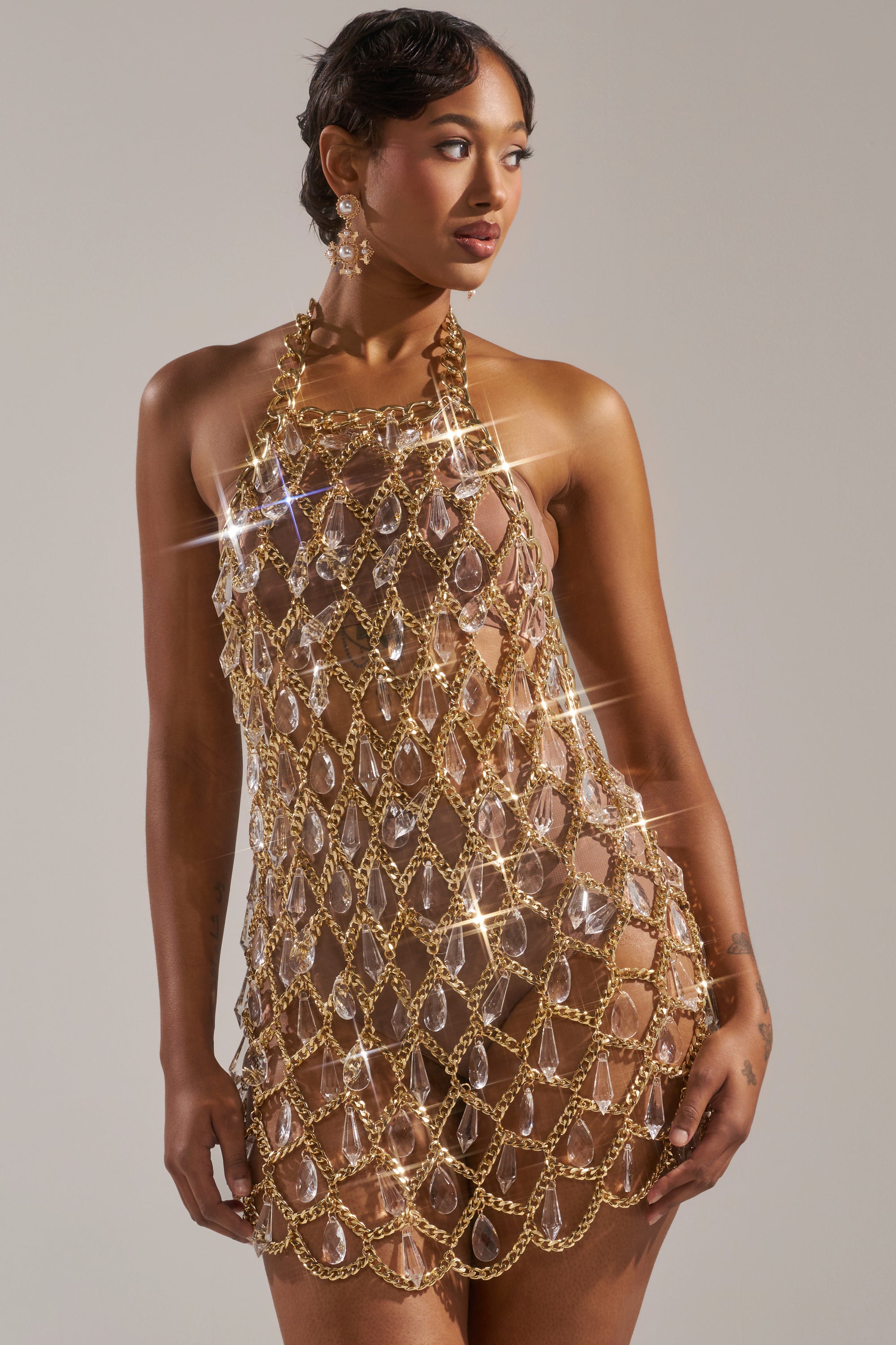 A woman poses confidently in the OH HOT DAMN RHINESTONE MINI DRESS, a transparent mini adorned with large gold chains and sparkling gem-like accents, paired with matching earrings for chic allure.