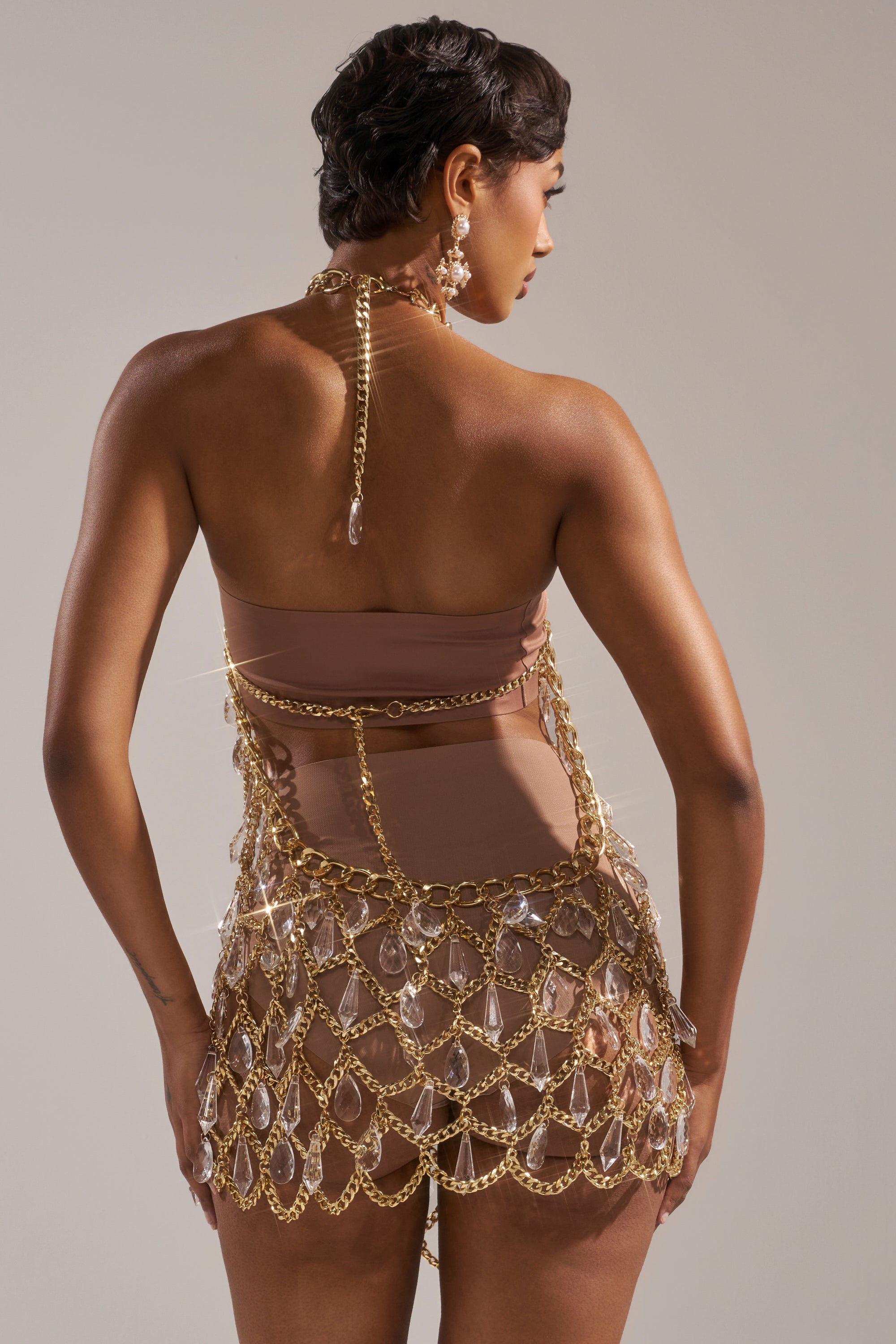 A person with short dark hair faces away, wearing the OH HOT DAMN RHINESTONE MINI DRESS—a nude bodysuit topped with a gold chain-link design and clear gemstones—plus dangling earrings, set against a plain background.