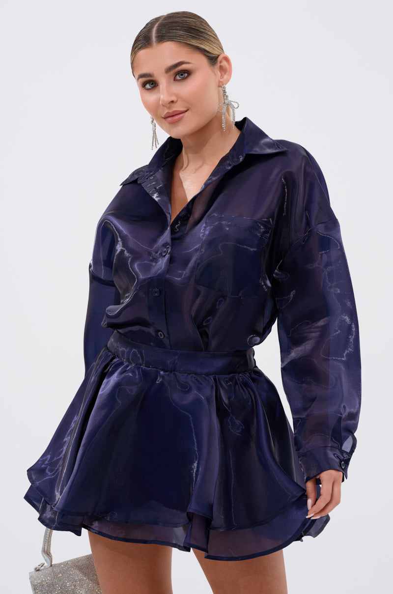 A woman models the CRAZY IN LOVE BUTTON DOWN BLOUSE in silky deep purple, paired with a matching flared mini skirt and dangling earrings, posing against a plain white background with her hair slicked back. 0