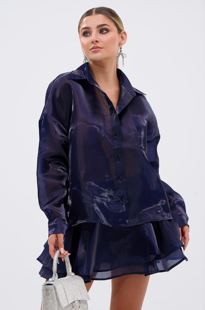 She wears the CRAZY IN LOVE BUTTON DOWN BLOUSE in silky navy with a matching ruffled skirt, holding a small silver glitter handbag. Slicked-back hair, statement earrings, and neutral makeup complete her chic look against a plain background. 1