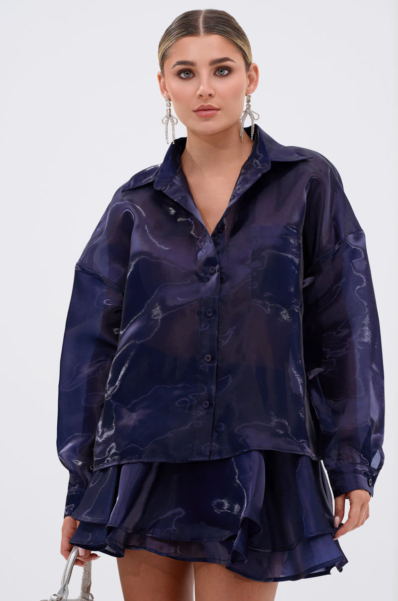A woman wears the CRAZY IN LOVE BUTTON DOWN BLOUSE, a silky navy long-sleeve top paired with a matching ruffled skirt, holding a small handbag and standing against a plain white background. 2