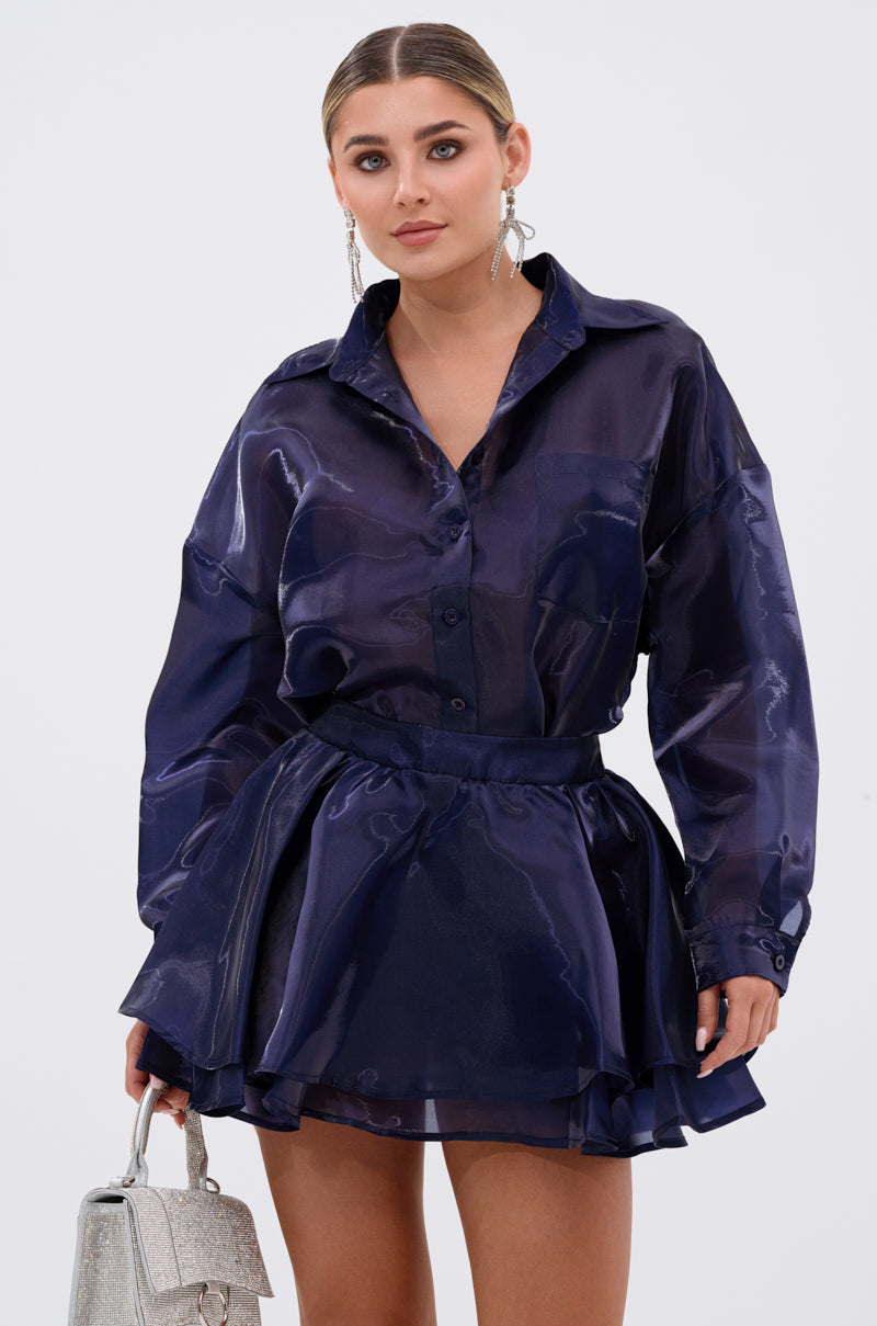 A woman wears the CRAZY IN LOVE BUTTON DOWN BLOUSE in silky navy with a matching flared skirt, holding a silver textured handbag. She has slicked-back hair, bold eye makeup, and statement earrings, standing against a white background. 3