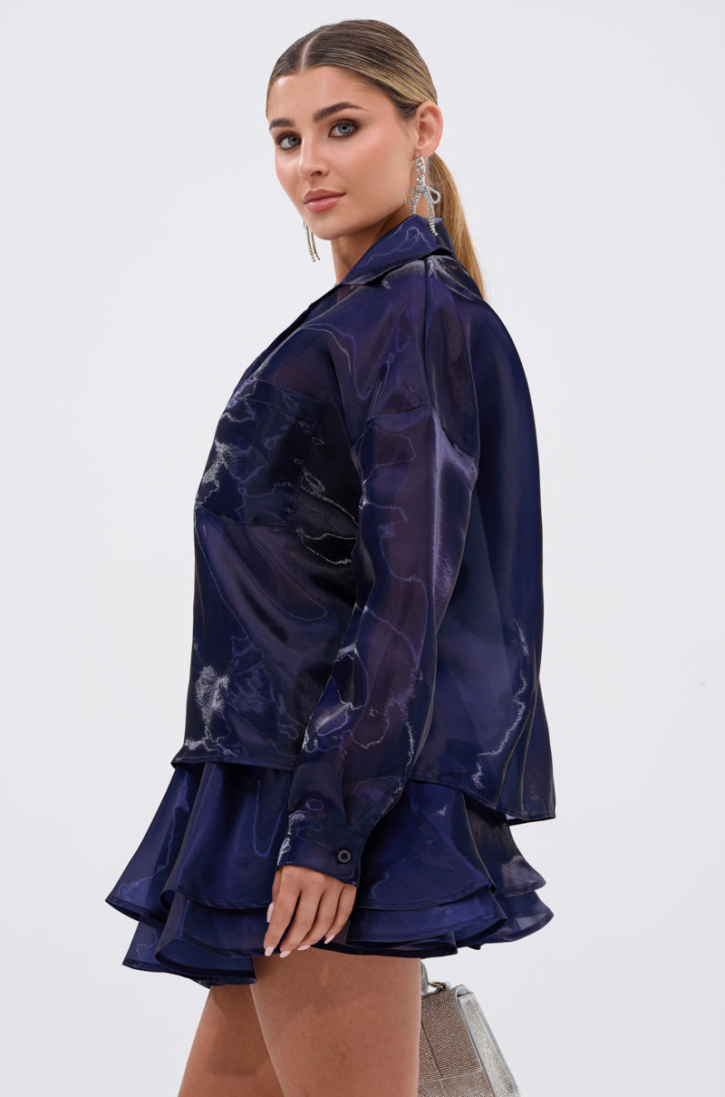 A woman with slicked-back hair wears the CRAZY IN LOVE BUTTON DOWN BLOUSE in silky navy, paired with a matching ruffled skirt. She holds a sparkly silver handbag and poses confidently against a plain light background. 4