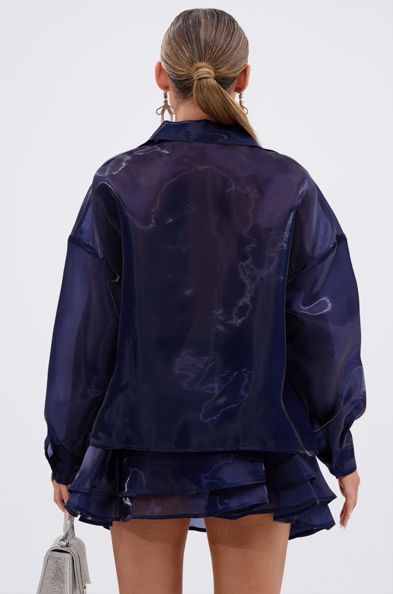 A woman with slicked-back hair is seen from behind in the silky, navy CRAZY IN LOVE BUTTON DOWN BLOUSE and a matching ruffled mini skirt, holding a silver handbag and wearing hoop earrings. 5