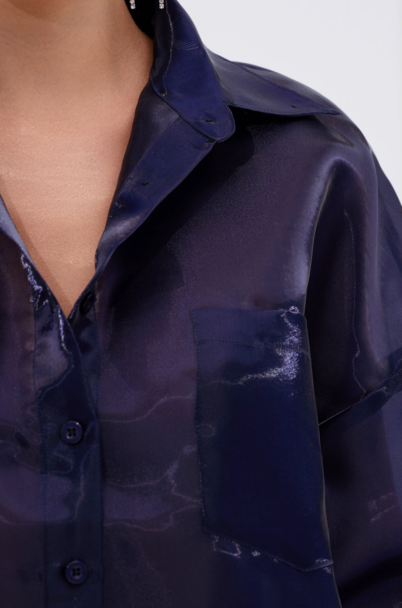 Close-up of a person wearing the CRAZY IN LOVE BUTTON DOWN BLOUSE, a silky navy, semi-sheer top with a collar and chest pocket. The glossy finish catches the light, and part of the person's neck and face is visible. 7