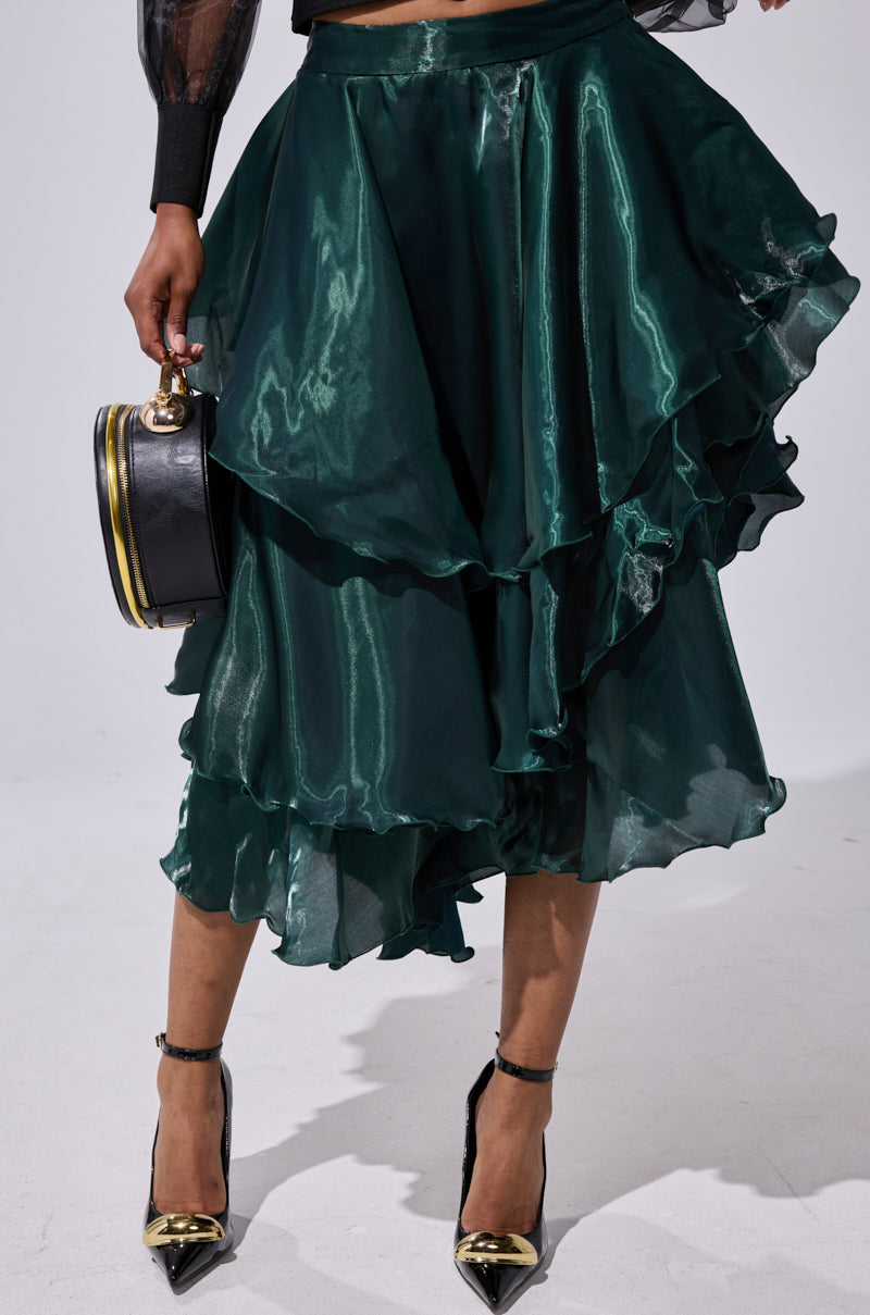 A person wearing the LUXE LIFE SATIN MIDI SKIRT in shiny dark green, paired with a black sheer top, black pointed heels with gold accents, and holding a round black handbag. 0