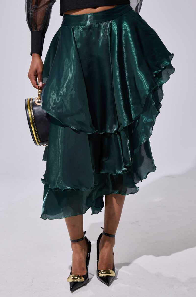 A person wears the LUXE LIFE SATIN MIDI SKIRT in shiny dark green with tiered ruffles, paired with a black sheer long-sleeve top, black pointed-toe heels with gold detail, and a small round black handbag. 1