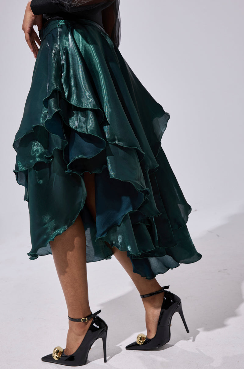A person wearing the LUXE LIFE SATIN MIDI SKIRT—a shiny, dark green, high-waisted tiered ruffle skirt with an uneven hem—paired with black heels featuring gold accents, standing against a plain light background. 2