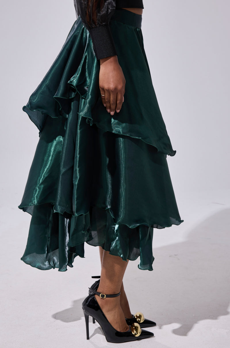 A person wearing the LUXE LIFE SATIN MIDI SKIRT in dark green with an asymmetrical hem and high waist, paired with black heels featuring gold accents, stands against a plain light background. 3
