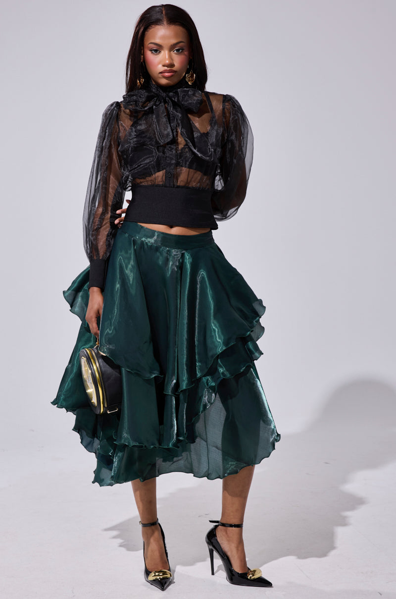 A woman models the LUXE LIFE SATIN MIDI SKIRT, paired with a sheer black blouse and bow. She accessorizes with a small round purse and black heels with gold accents against a simple, light backdrop. 5