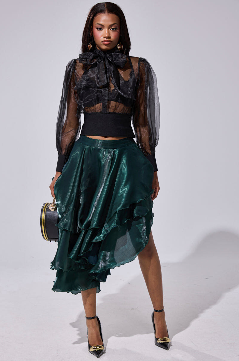 A woman stands confidently in a sheer black blouse with a large bow and long sleeves, paired with the LUXE LIFE SATIN MIDI SKIRT in dark green. She holds a small handbag and wears black heels. 6