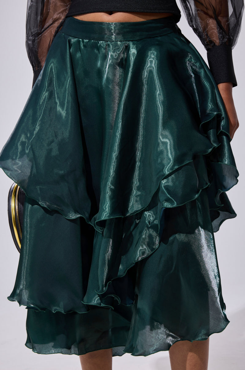 Wearing the LUXE LIFE SATIN MIDI SKIRT in shiny dark green with ruffled tiers, the person pairs this high-waisted, mid-length skirt with a small round black clutch. Only their lower torso and part of their arms are visible. 7