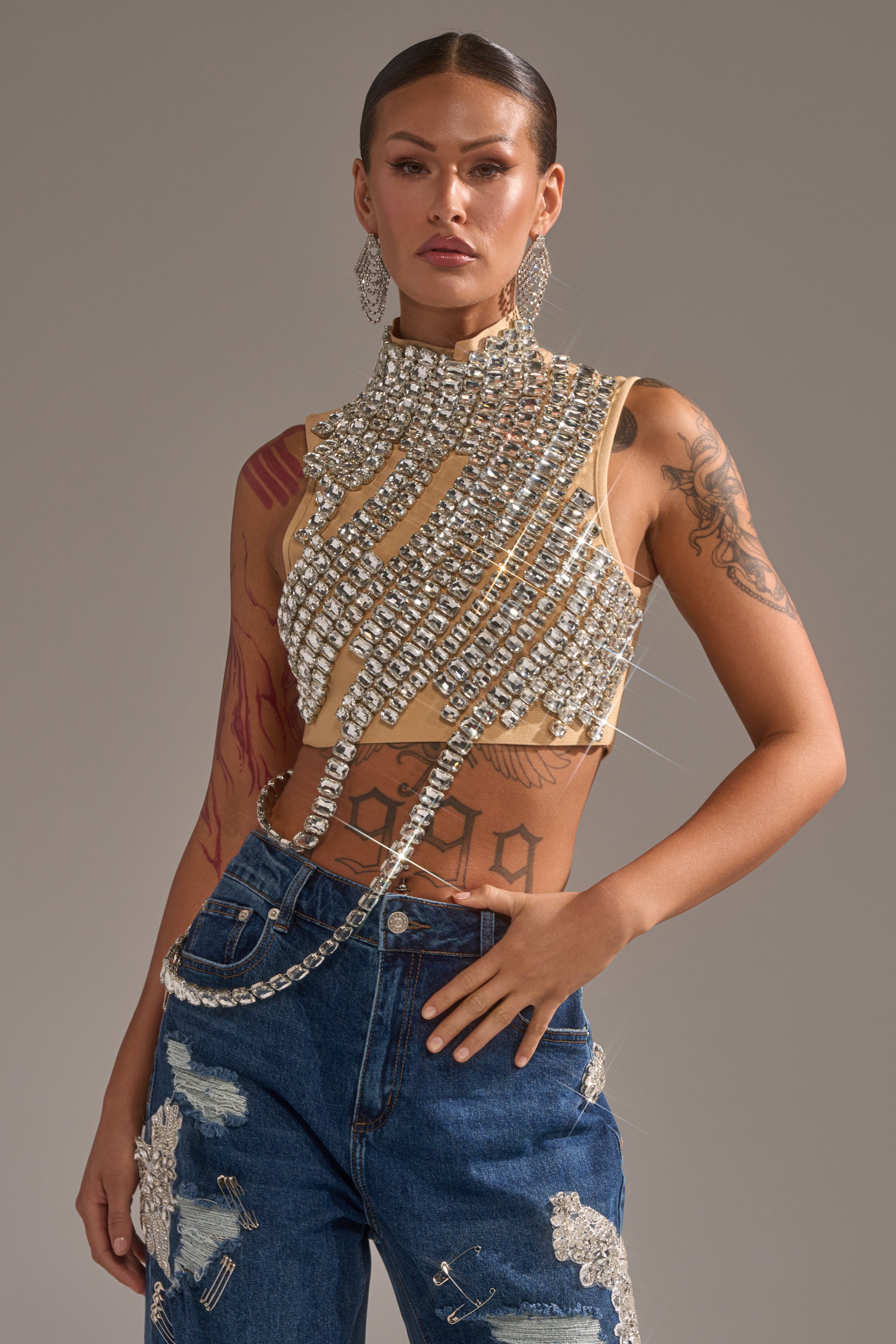 A tattooed woman poses confidently in the ITS YOUR TIME RHINESTONE MOCK NECK BLOUSE and blue embellished jeans, accessorized with oversized rhinestone earrings and slicked-back hair.