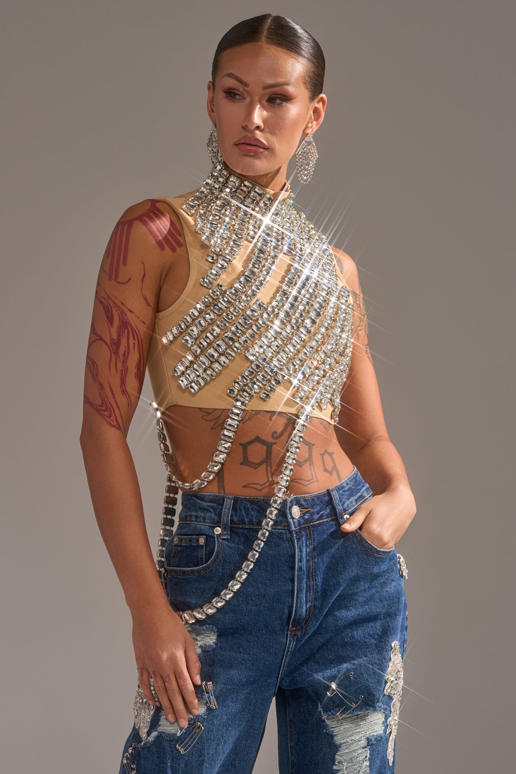 A person with slicked-back hair wears the ITS YOUR TIME RHINESTONE MOCK NECK BLOUSE and high-waisted blue jeans adorned with rhinestone patches, styled with visible tattoos and large, dangling earrings for a bold, sparkling look.