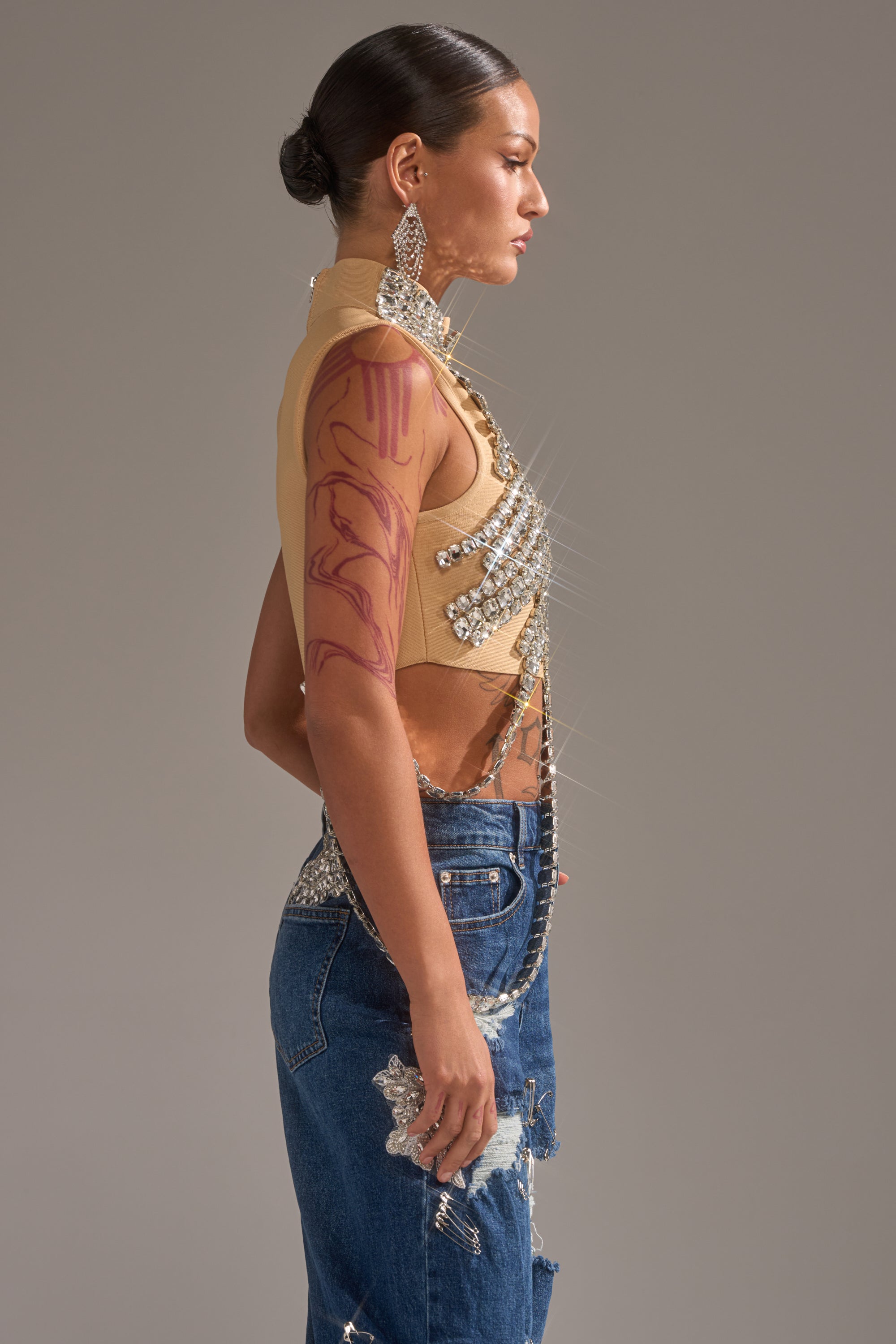 A person in profile wears the ITS YOUR TIME RHINESTONE MOCK NECK BLOUSE, styled with blue jeans featuring glitter embellishments, large dangling earrings, and a red upper arm tattoo, standing before a plain background.