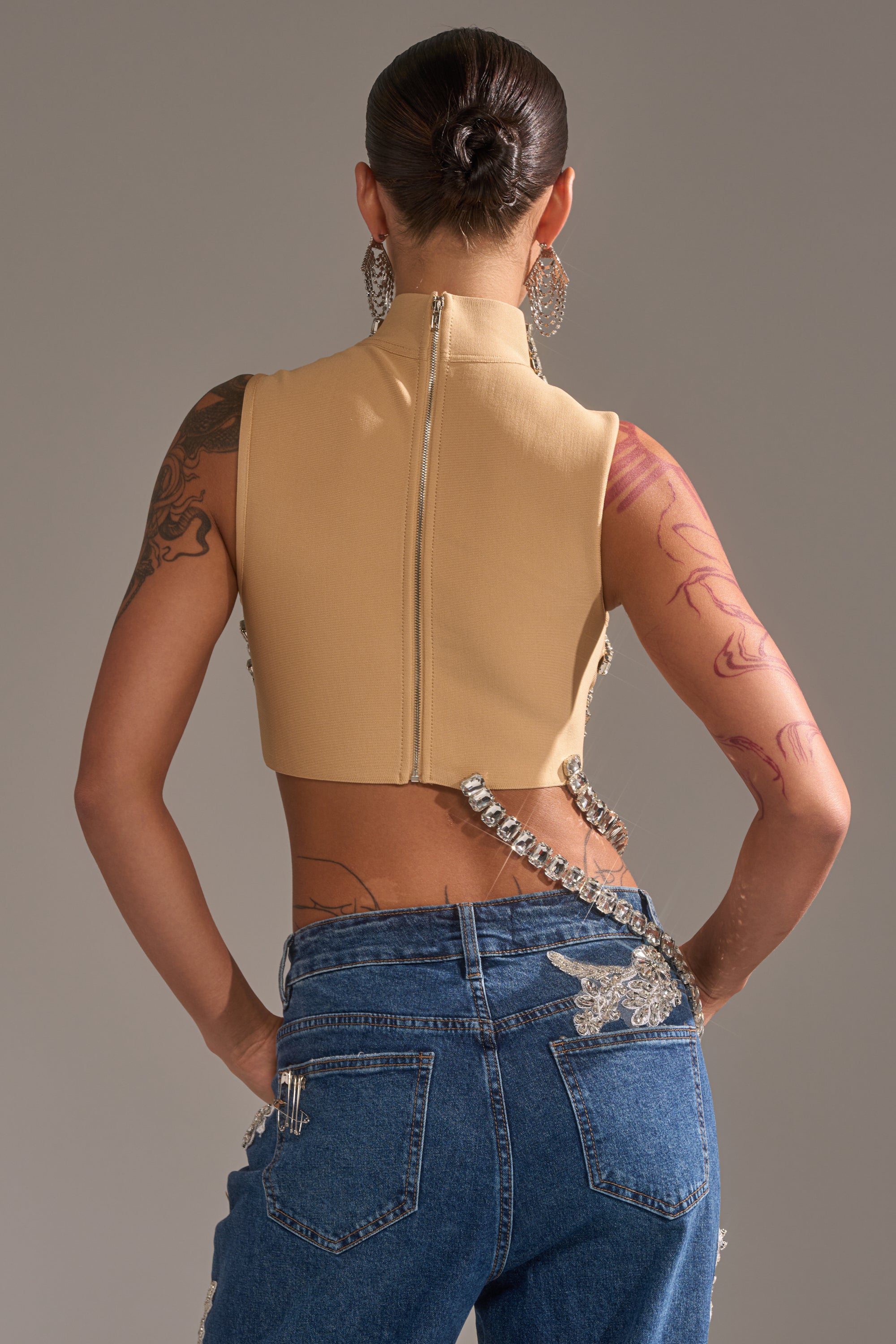 A person with their hair in a bun models the AKIRA Label ITS YOUR TIME RHINESTONE MOCK NECK BLOUSE with blue jeans featuring silver embellishments. Visible tattoos cover their left arm and right forearm.