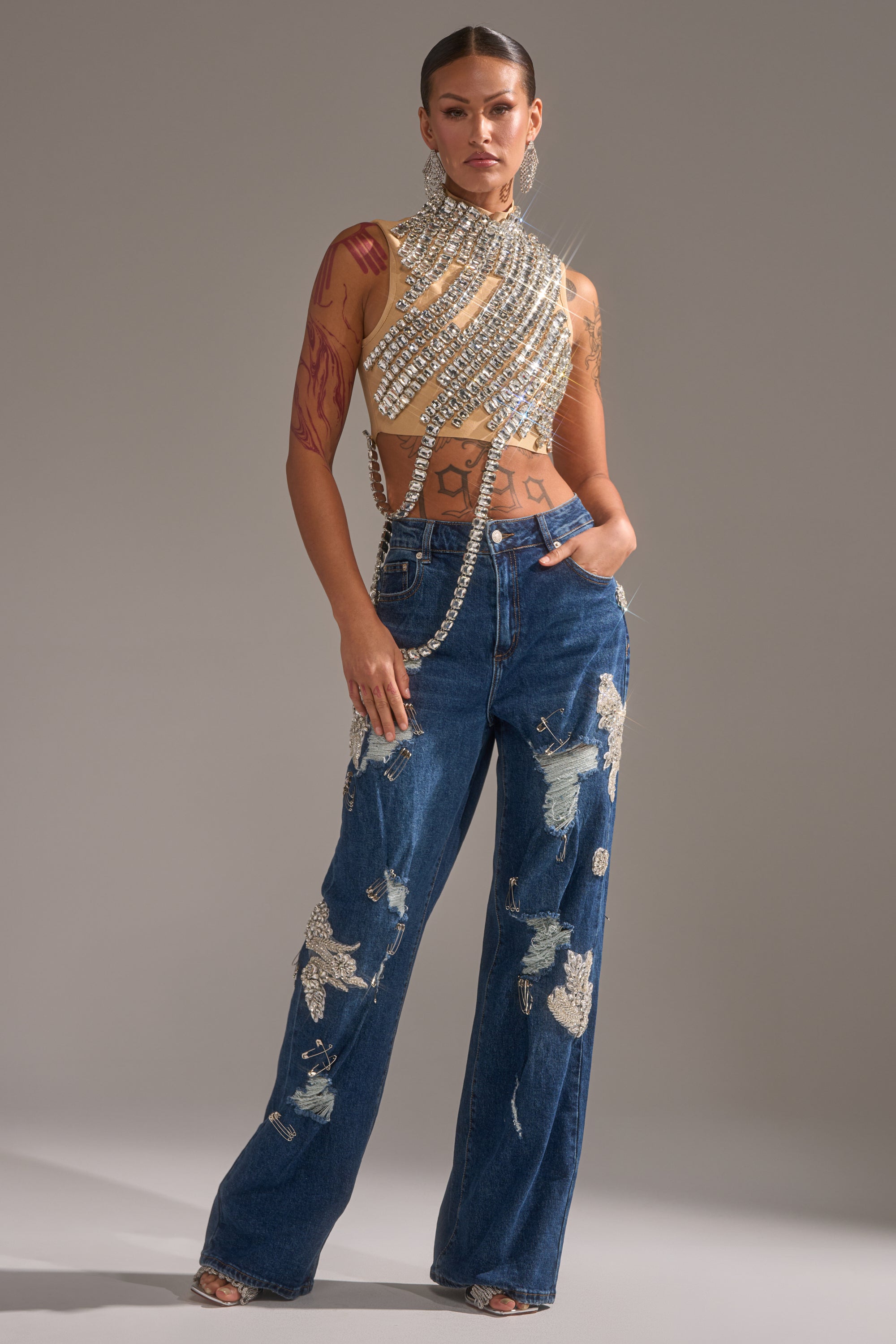 A person poses confidently in the ITS YOUR TIME RHINESTONE MOCK NECK BLOUSE and blue glittering baggy jeans, showcasing tattoos, slicked-back hair, and large earrings against a plain gray background.