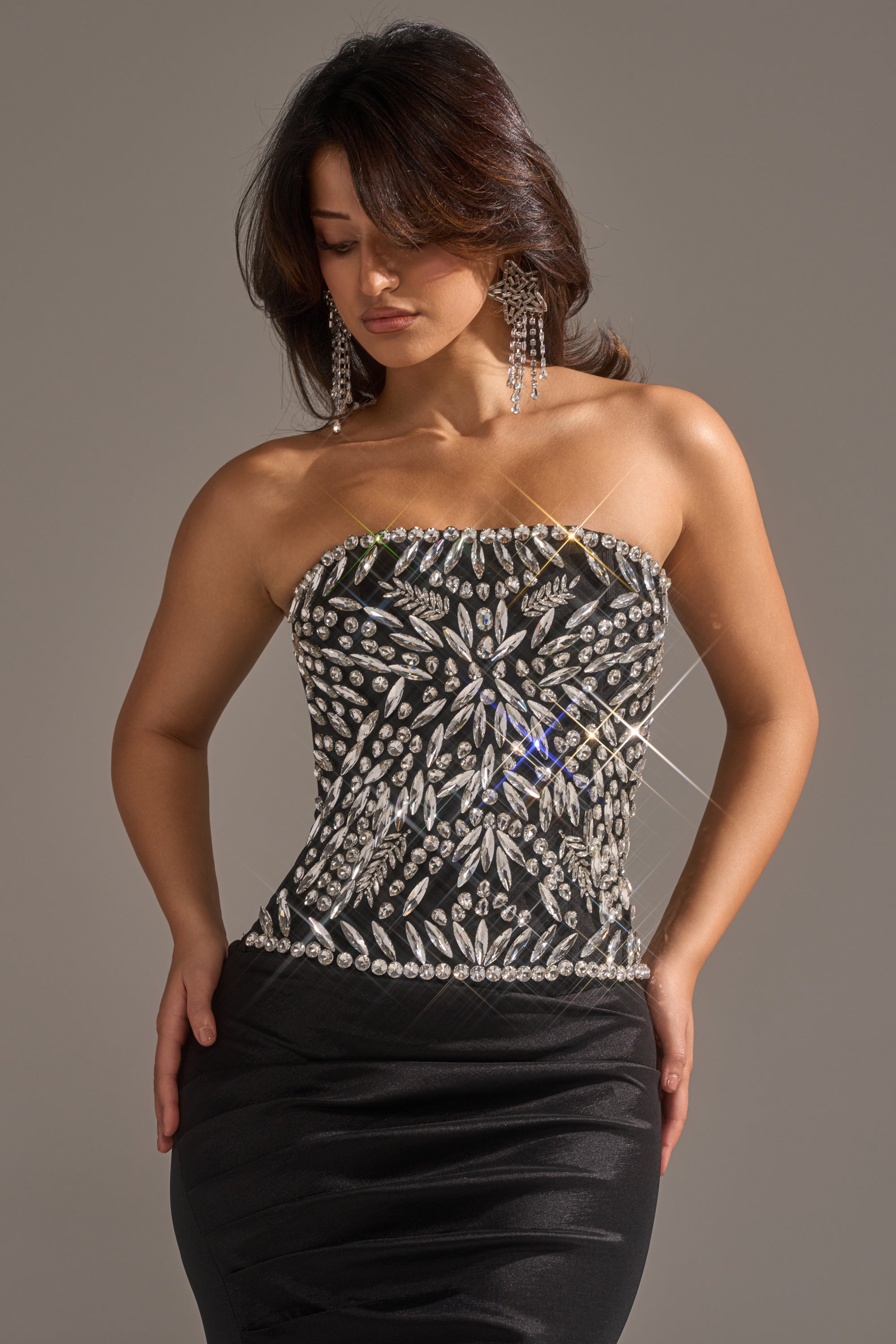 A woman in the QUEEN OF THE NIGHT RHINESTONE CORSET, a strapless black dress with gemstone accents, stands against a plain backdrop, looking down with her hands on her hips. She has long dark hair and wears large dangling earrings. 0