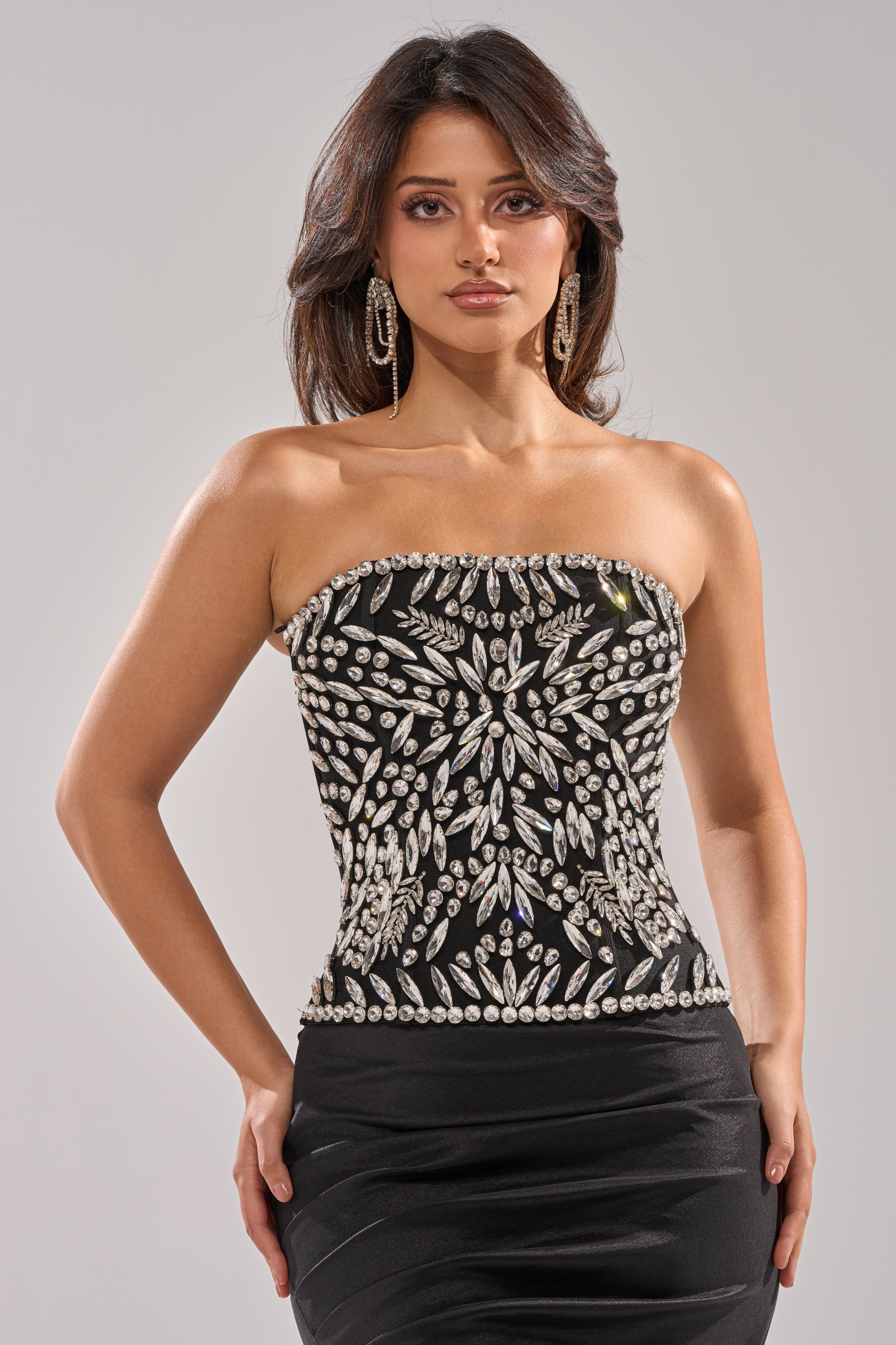 A woman with long dark hair wears the QUEEN OF THE NIGHT RHINESTONE CORSET— a strapless black dress with intricate silver beading—paired with large hoop earrings, standing against a plain light gray background. 1