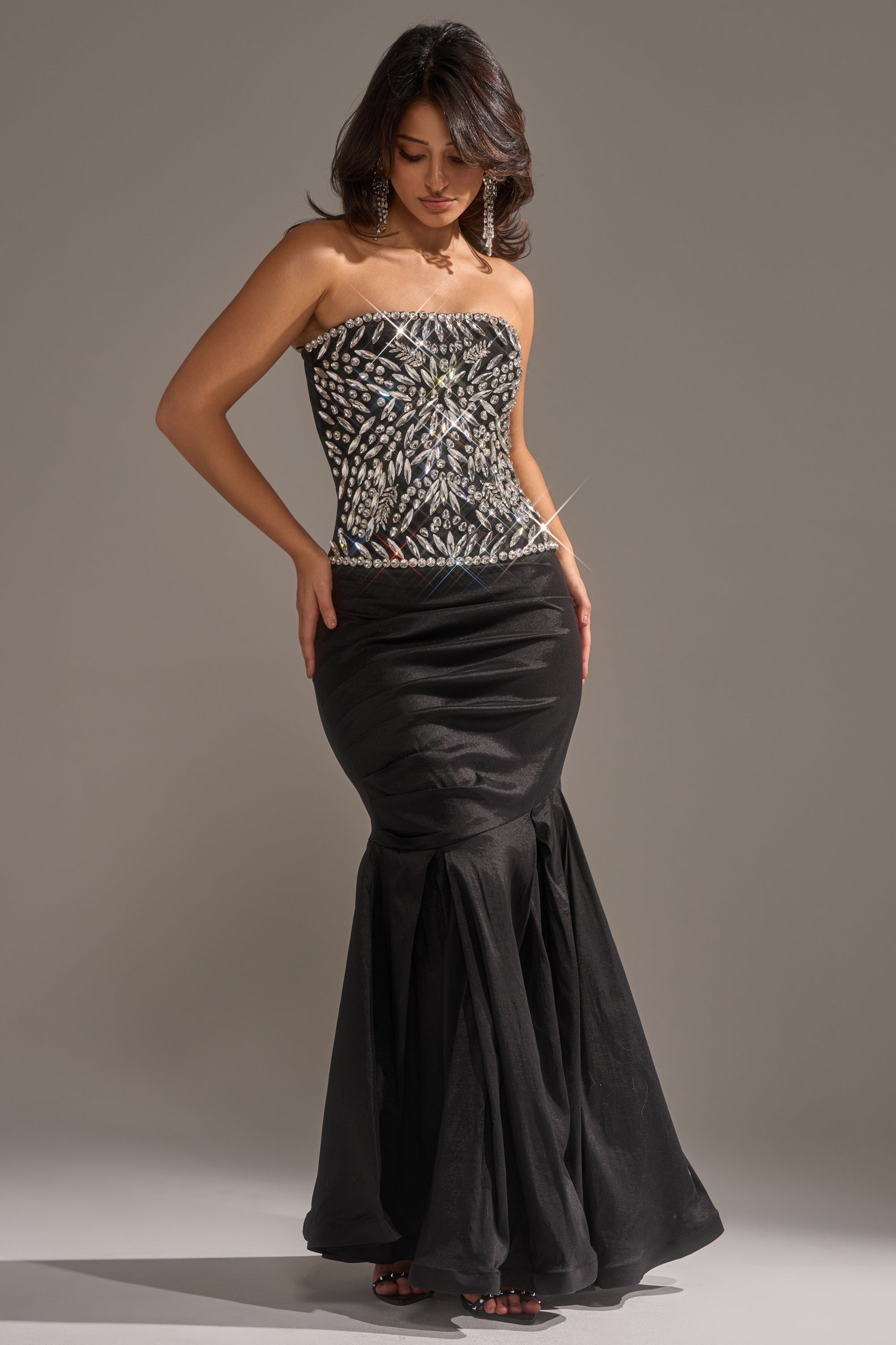 A woman wearing the QUEEN OF THE NIGHT RHINESTONE CORSET black evening gown with a strapless neckline and rhinestone bodice stands against a plain backdrop, looking down with one hand on her hip and the other on her thigh. 4