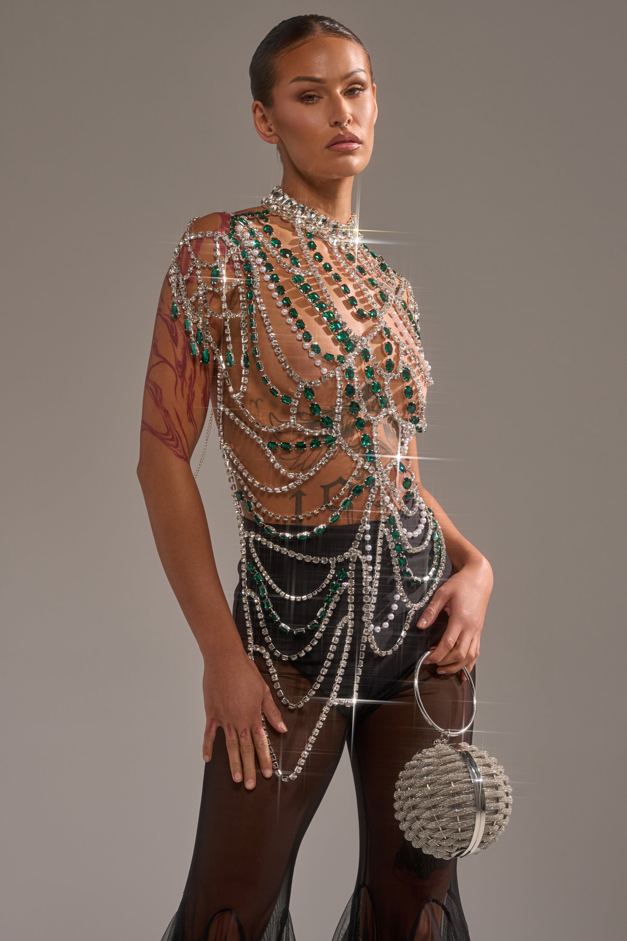 Wearing the DANCING QUEEN RHINESTONE CHAIN TOP with sparkling green and silver gems, black sheer pants, and a round silver beaded bag, they pose confidently with slicked-back hair and a serious expression for a bold, standout look. 0
