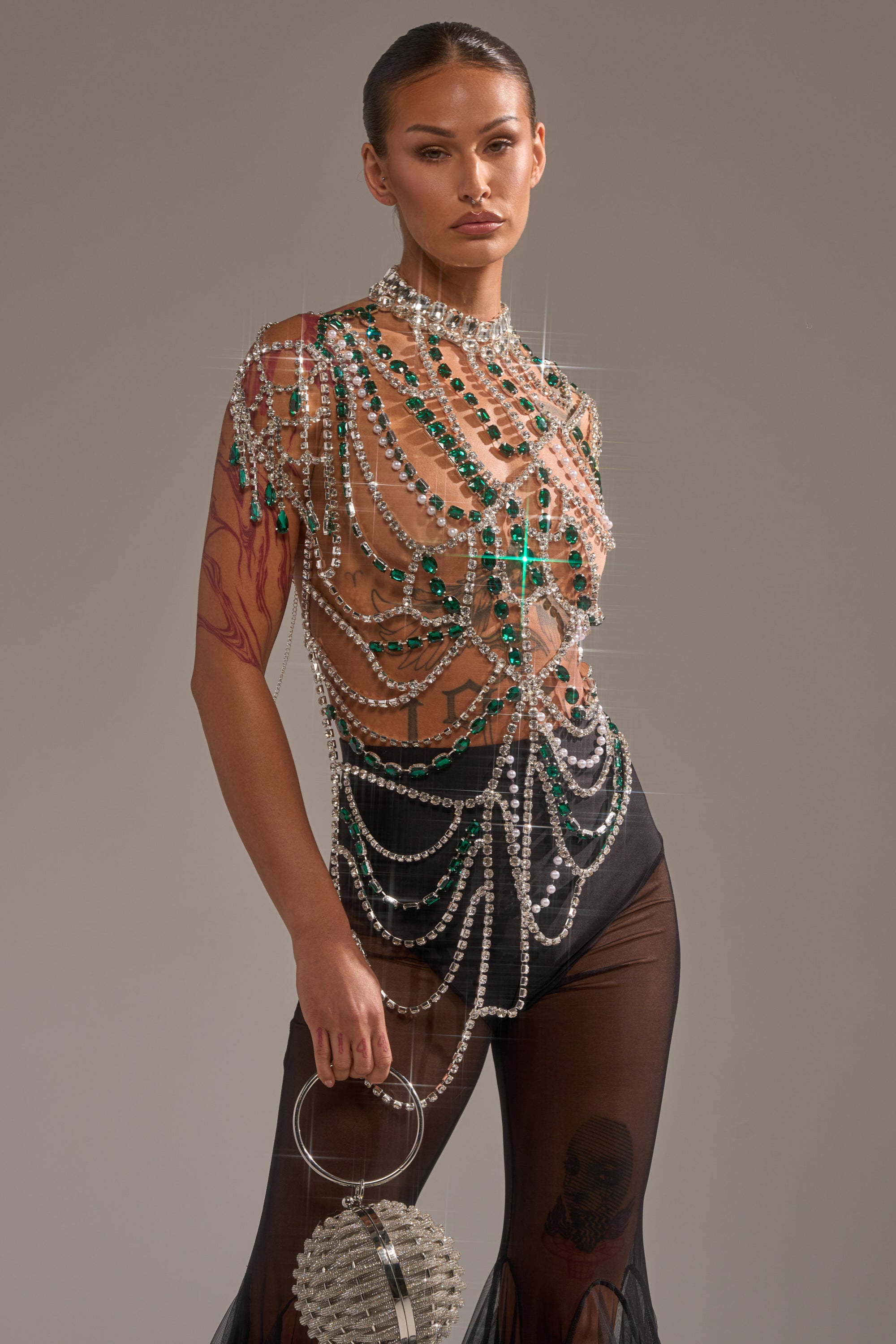 A person with slicked-back hair wears the DANCING QUEEN RHINESTONE CHAIN TOP adorned with gemstone detailing and green crystals, paired with black sheer pants. They hold a round silver handbag and reveal an arm tattoo against a neutral gray background. 1