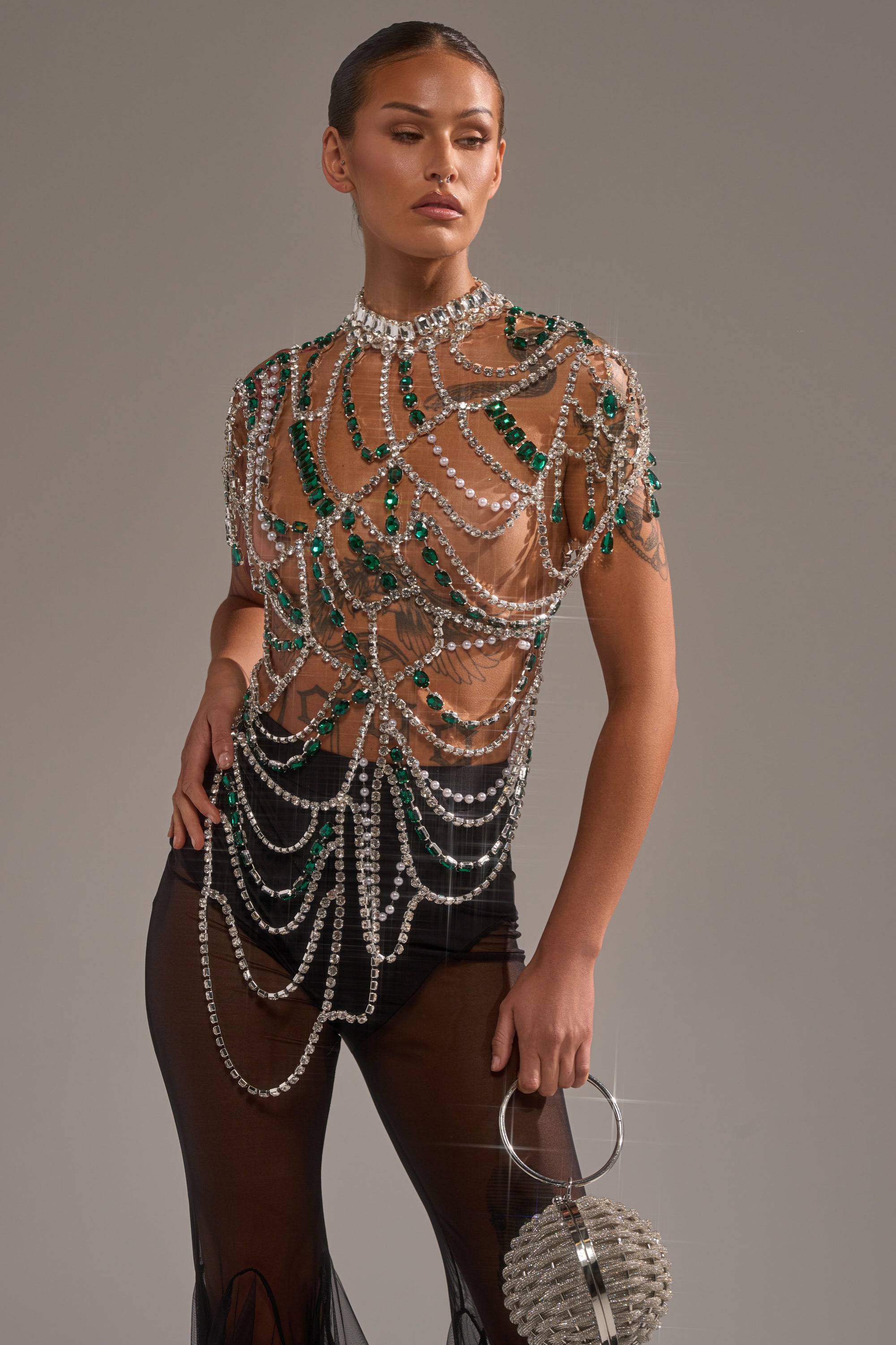 Wearing the DANCING QUEEN RHINESTONE CHAIN TOP, adorned with sparkling gemstones and draped chains, a person holds a round shiny handbag. The plain background highlights the top's stunning embellished details. 2