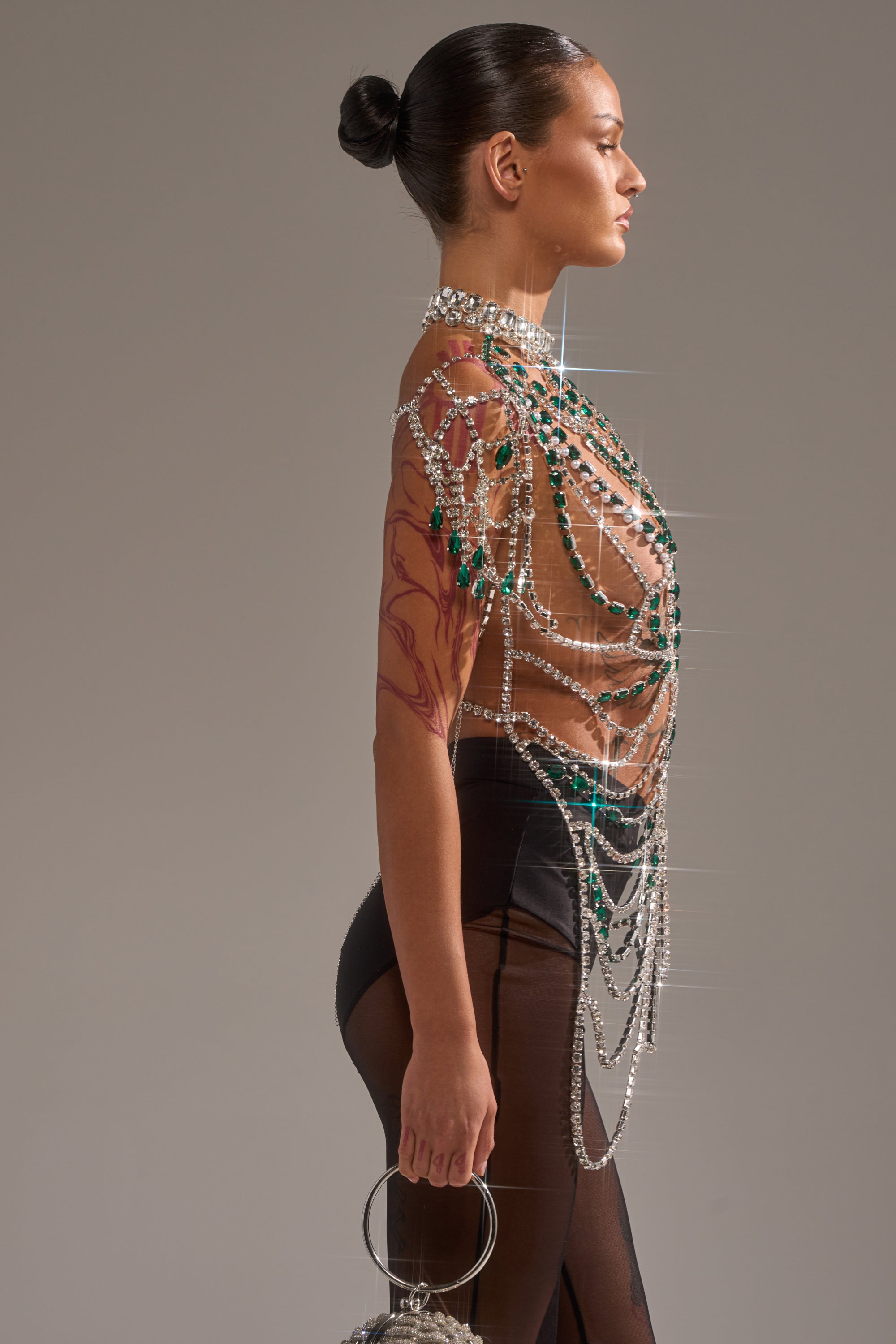 A woman in profile wears the DANCING QUEEN RHINESTONE CHAIN TOP with draped pearls and crystals, paired with a sheer black skirt. Her hair is in a sleek bun, she carries a small round handbag, and showcases a colorful tattoo on her arm. 3