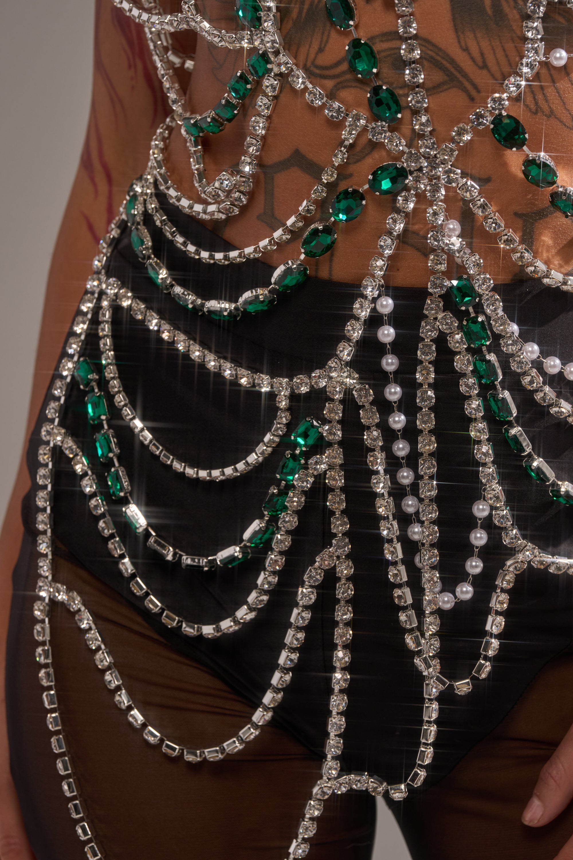 A close-up of a person wearing sheer black pants and the DANCING QUEEN RHINESTONE CHAIN TOP, adorned with rhinestones, green gems, and pearls along the hips and waist, revealing tattooed skin underneath. 6