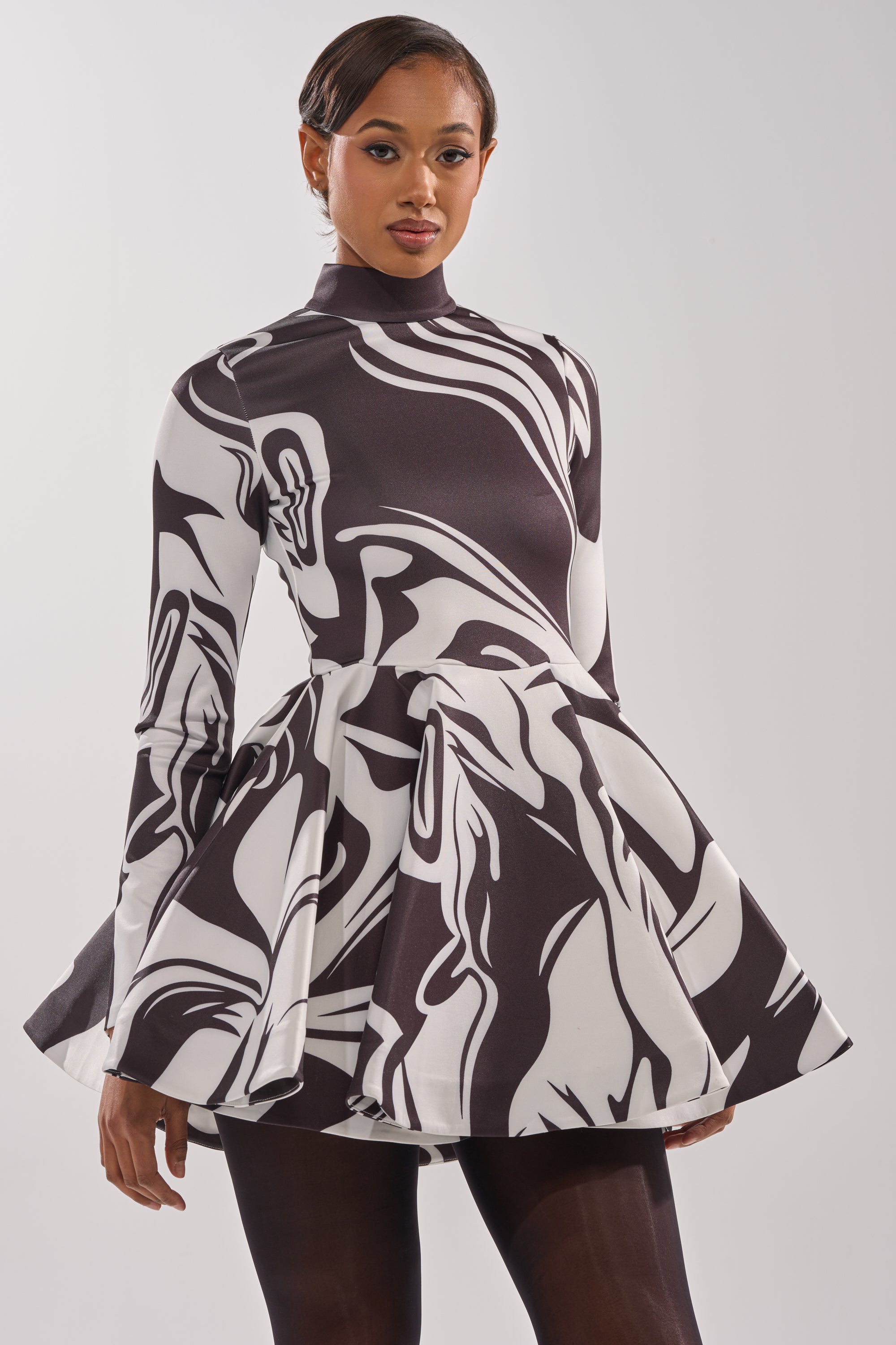 A woman models the GROOVY BABY MOCKNECK MINI DRESS, featuring a retro black and white swirl print, high neckline, long sleeves, and flared skirt against a plain white background.