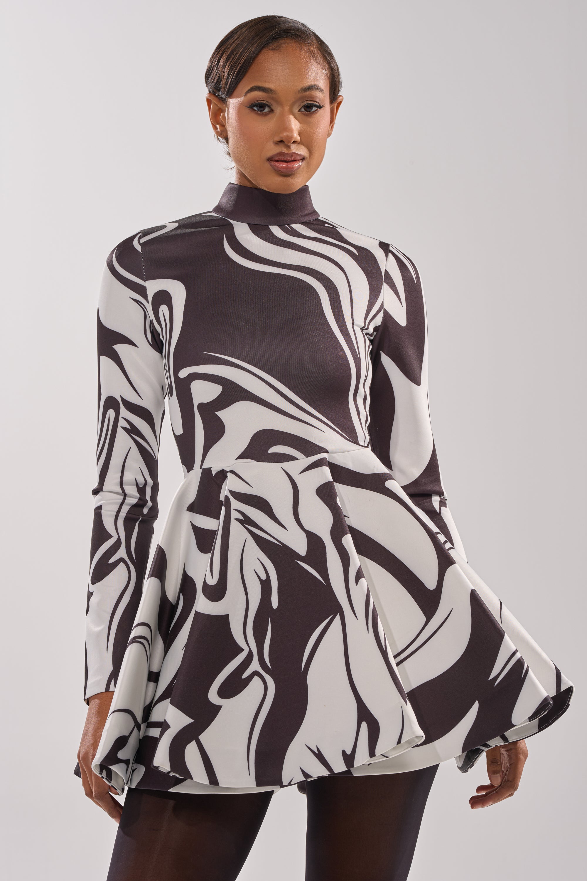 A woman models the GROOVY BABY MOCKNECK MINI DRESS, a retro-inspired black and white swirl design with long sleeves, high neckline, fitted bodice, and flared short skirt, standing against a plain white background.