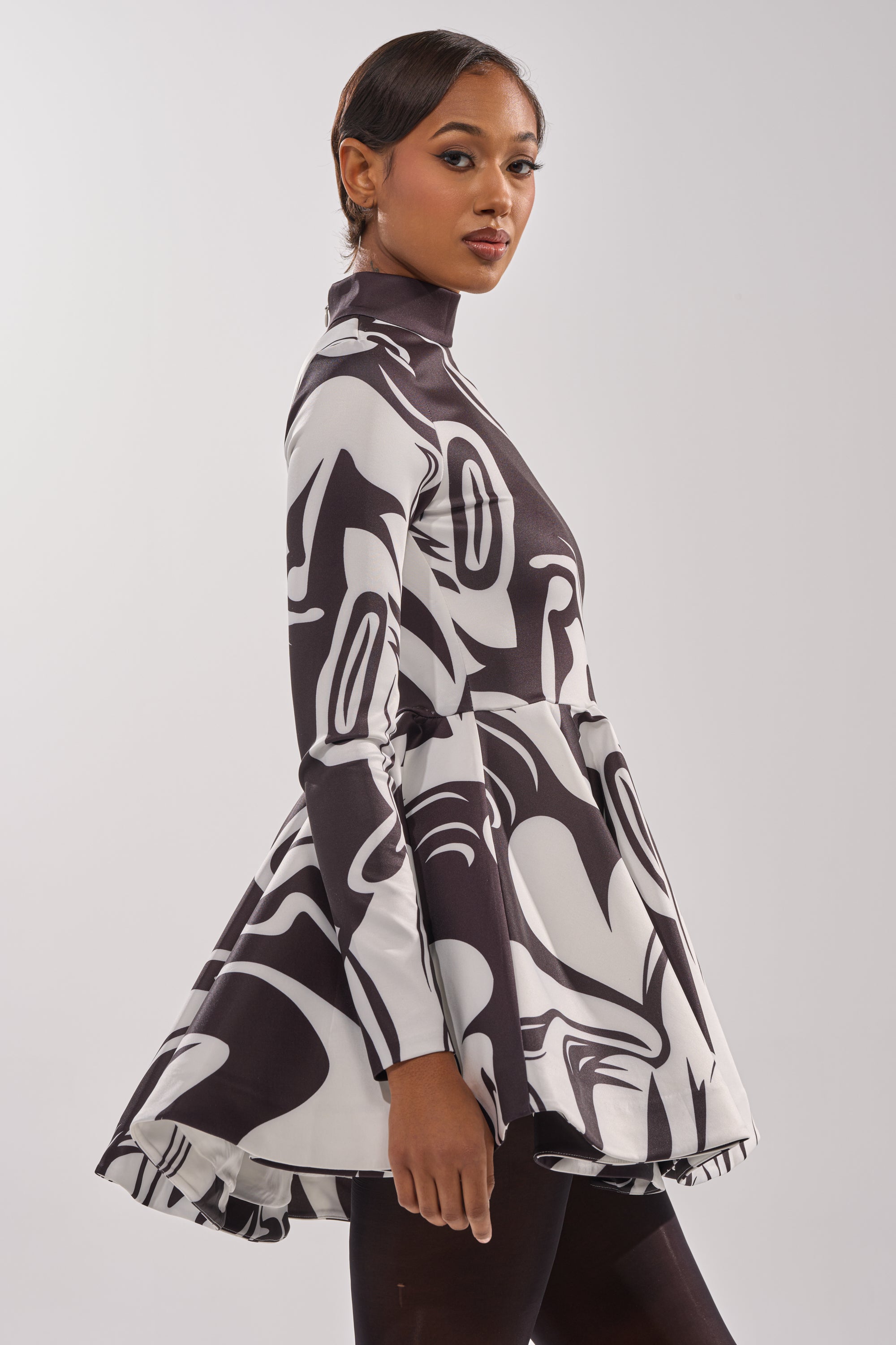A woman stands sideways, looking at the camera, in the GROOVY BABY MOCKNECK MINI DRESS—a retro black and white swirl mini dress with long sleeves, a high neck, flared skirt, and her hair styled neatly back.