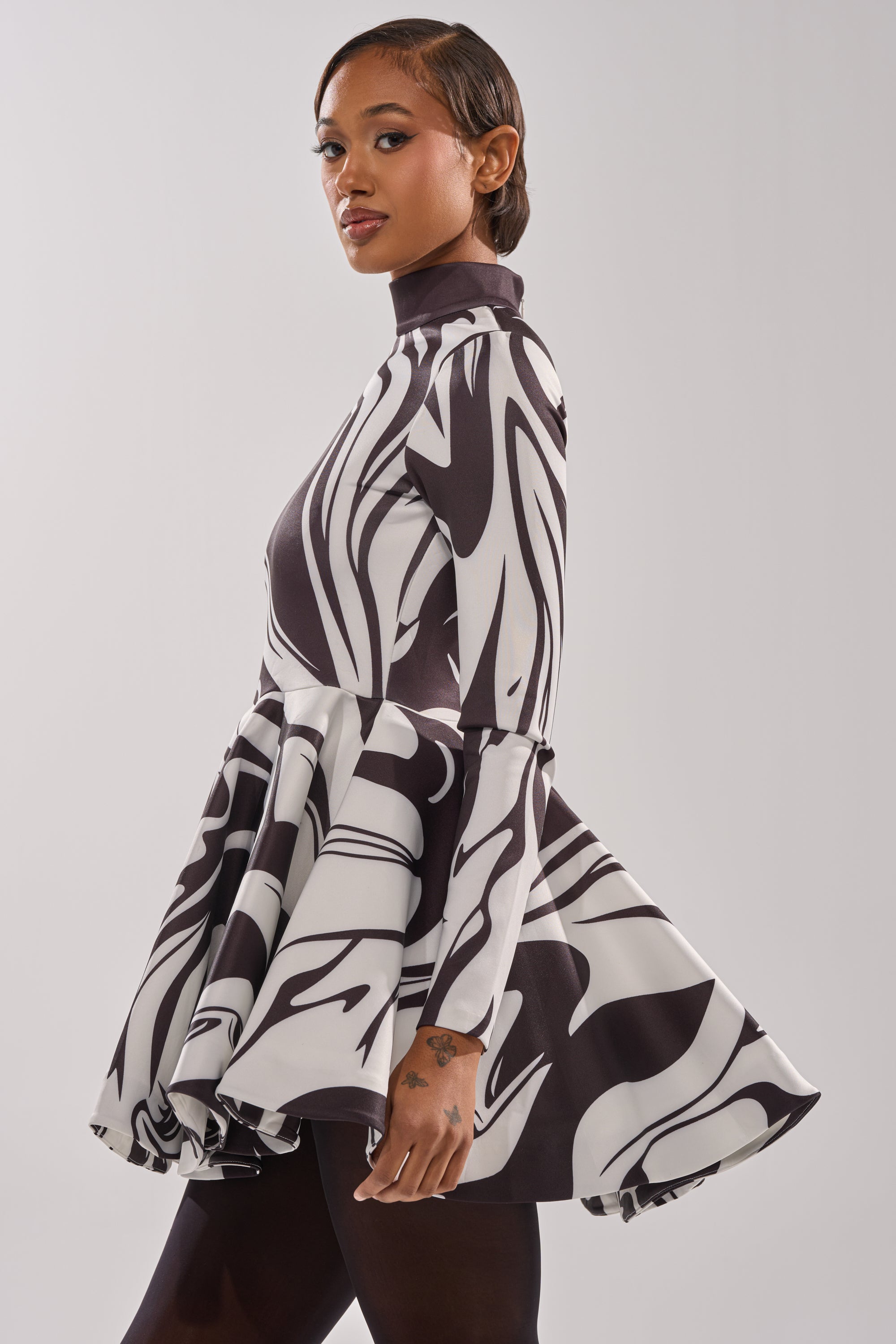 A woman with short hair and minimalist makeup poses confidently in the GROOVY BABY MOCKNECK MINI DRESS, featuring a black-and-white swirl print, retro vibe, high neck, and flared long sleeves, against a light background.