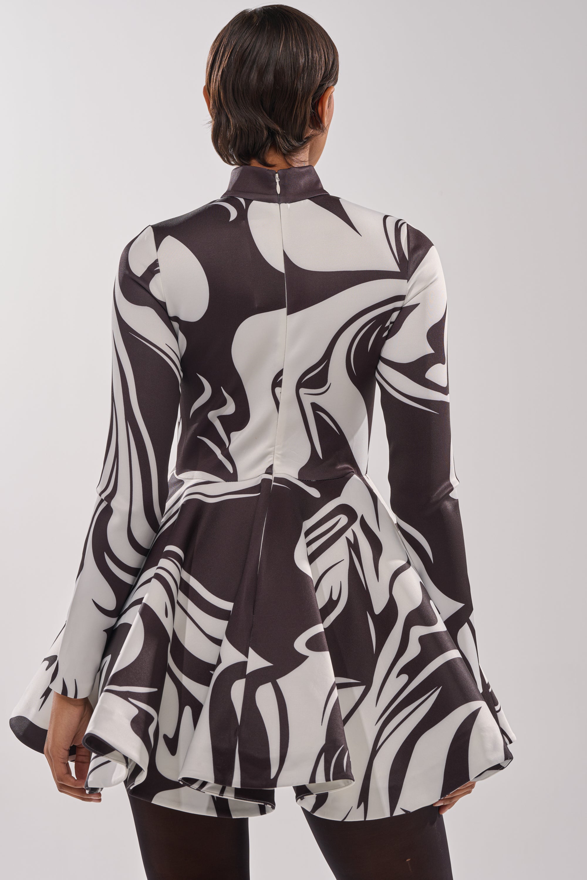 A person with short hair is seen from behind wearing the GROOVY BABY MOCKNECK MINI DRESS, featuring a black and white swirl print, long sleeves, flared skirt, and center zipper for a retro-inspired look.
