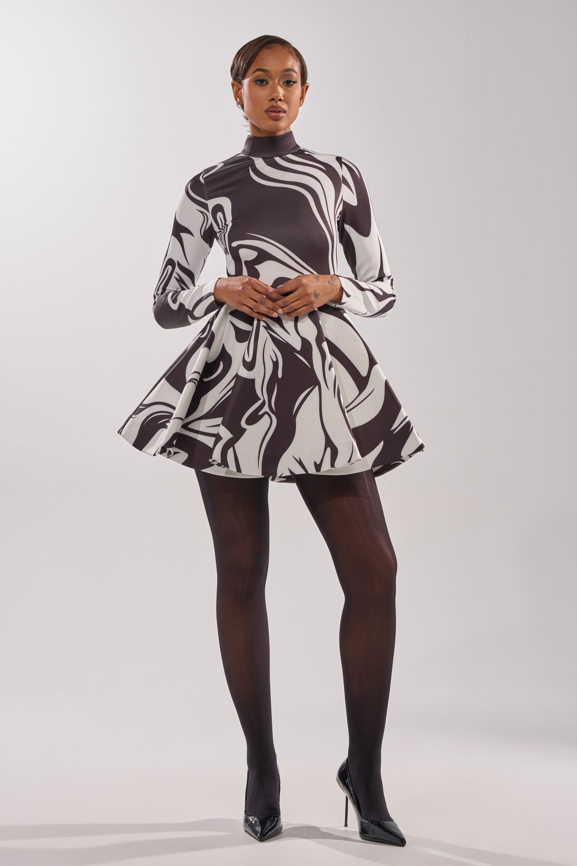 A woman stands confidently in the GROOVY BABY MOCKNECK MINI DRESS, featuring black and white retro-inspired swirls, styled with sheer black tights and heels against a plain white background.