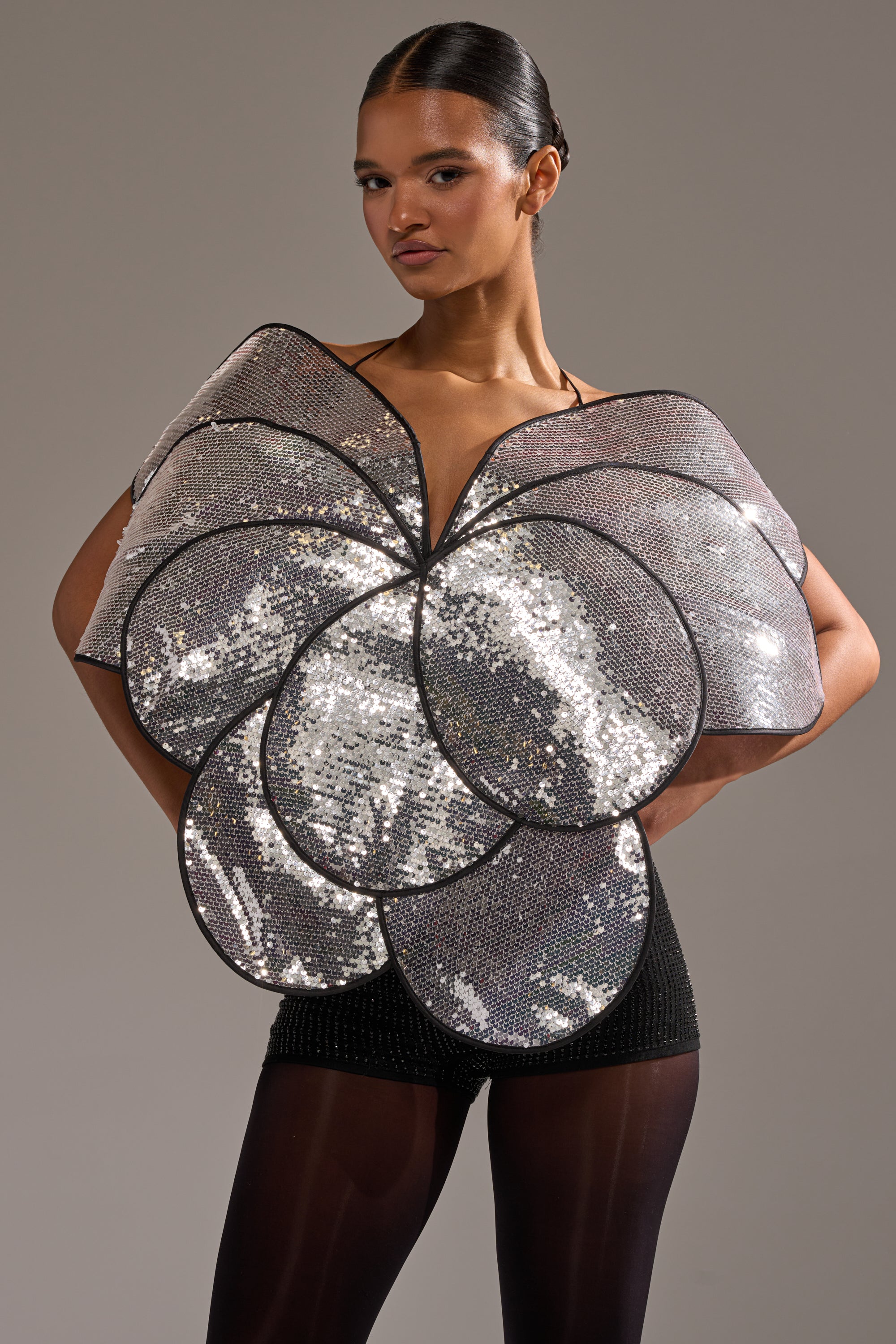 A woman poses confidently in the LA DIVINA SEQUIN TOP—an oversized, silver, flower petal-shaped top with sequins—paired with black shorts and sheer tights against a plain gray background. 0