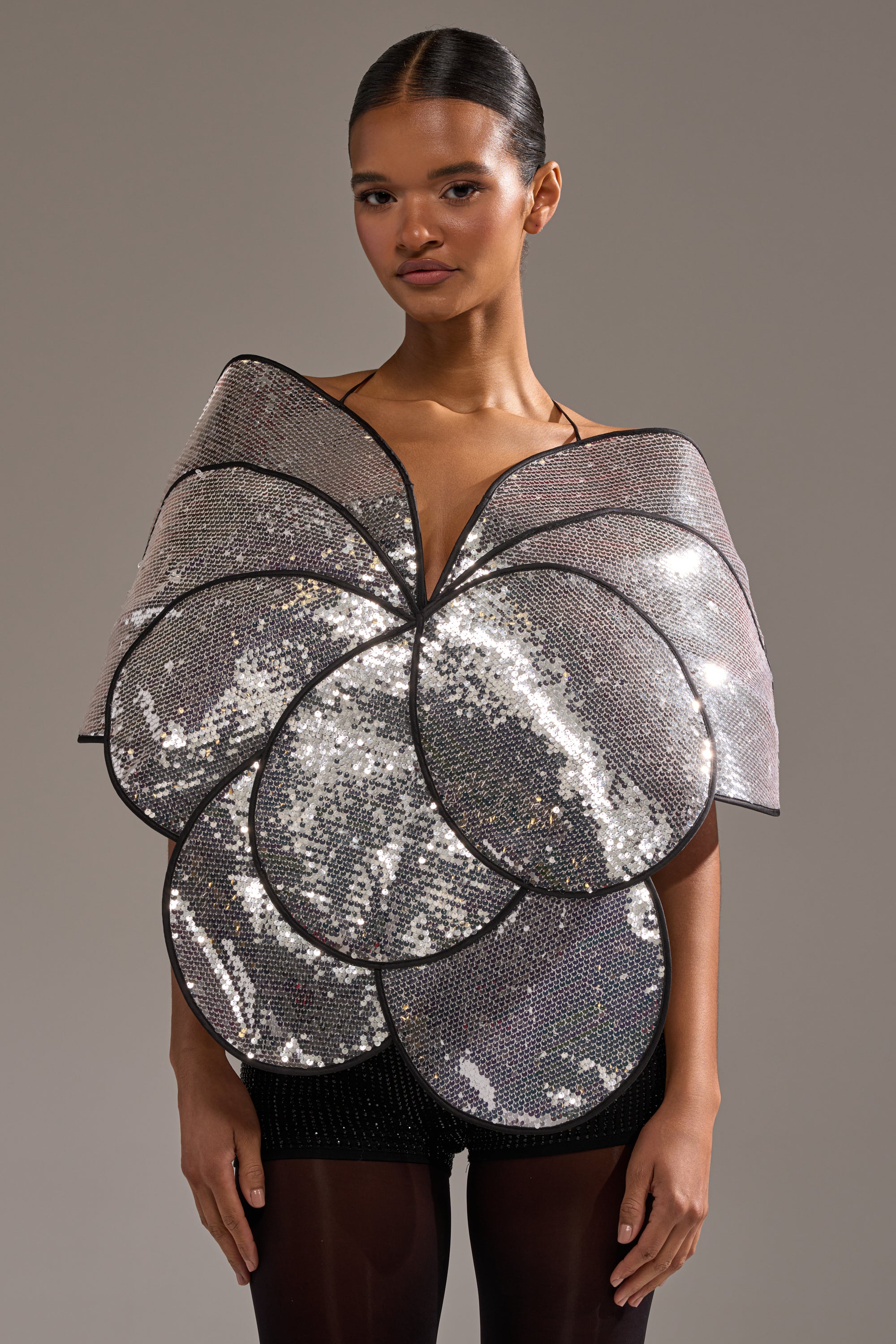 A woman models the LA DIVINA SEQUIN TOP, designed with large silver petal-shaped sequins enveloping her torso and shoulders, standing out beautifully against her simple hairstyle and a neutral background. 1