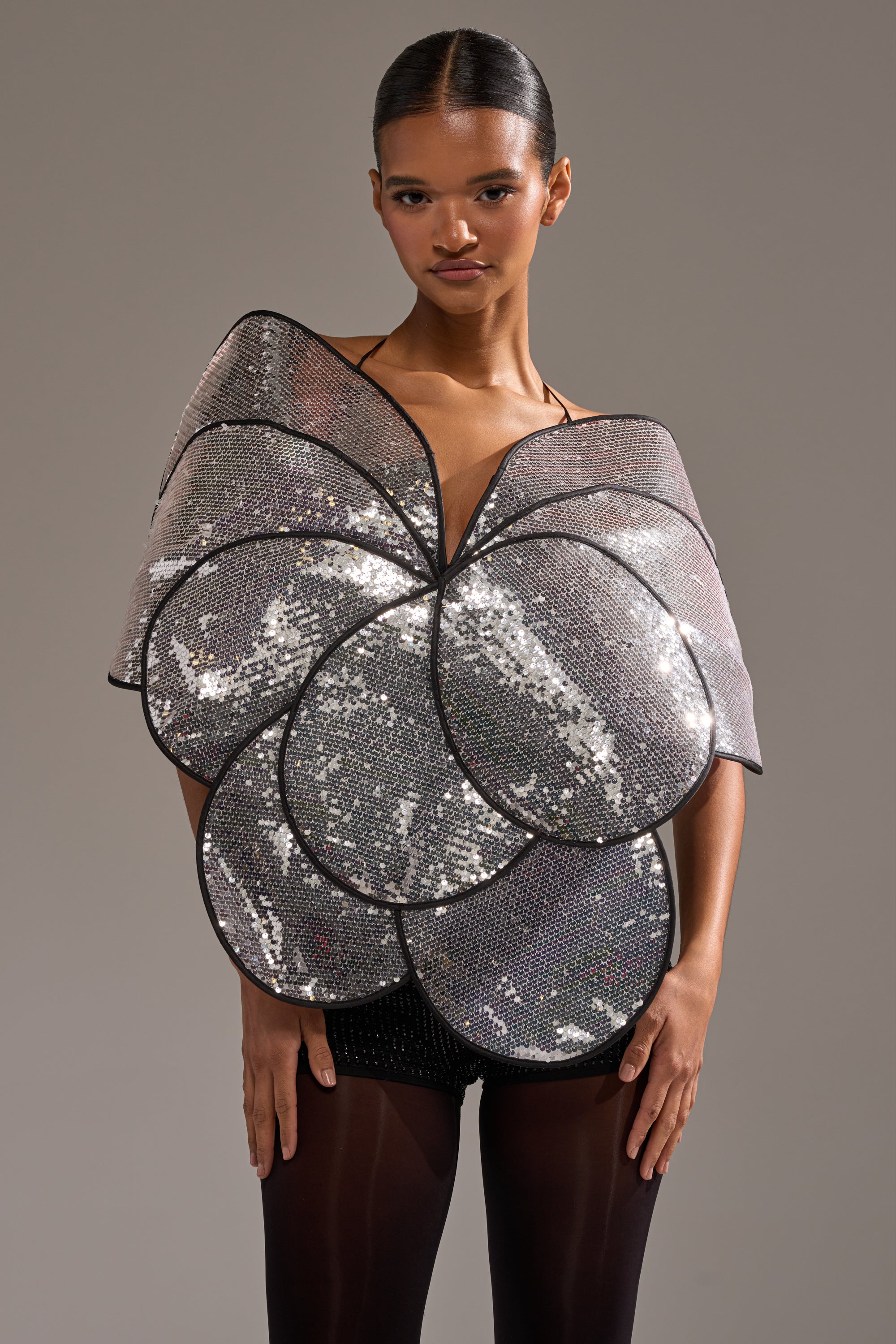 Someone wearing the LA DIVINA SEQUIN TOP, a shiny silver petal-shaped top with black outlines resembling a large flower, paired with black shorts and tights, stands against a plain background. 2