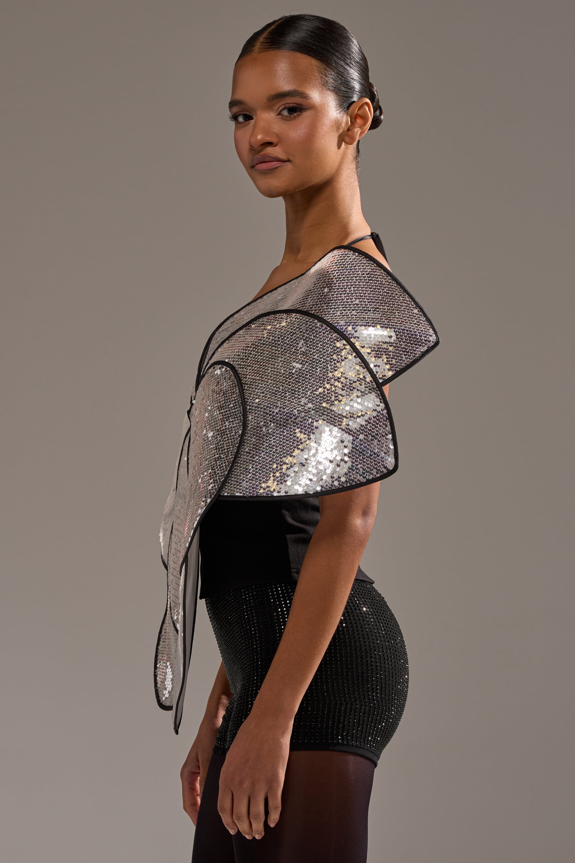 A woman poses in profile against a plain gray background, wearing the LA DIVINA SEQUIN TOP—an elegant black top with sparkling silver, wing-like shoulder embellishments. 3