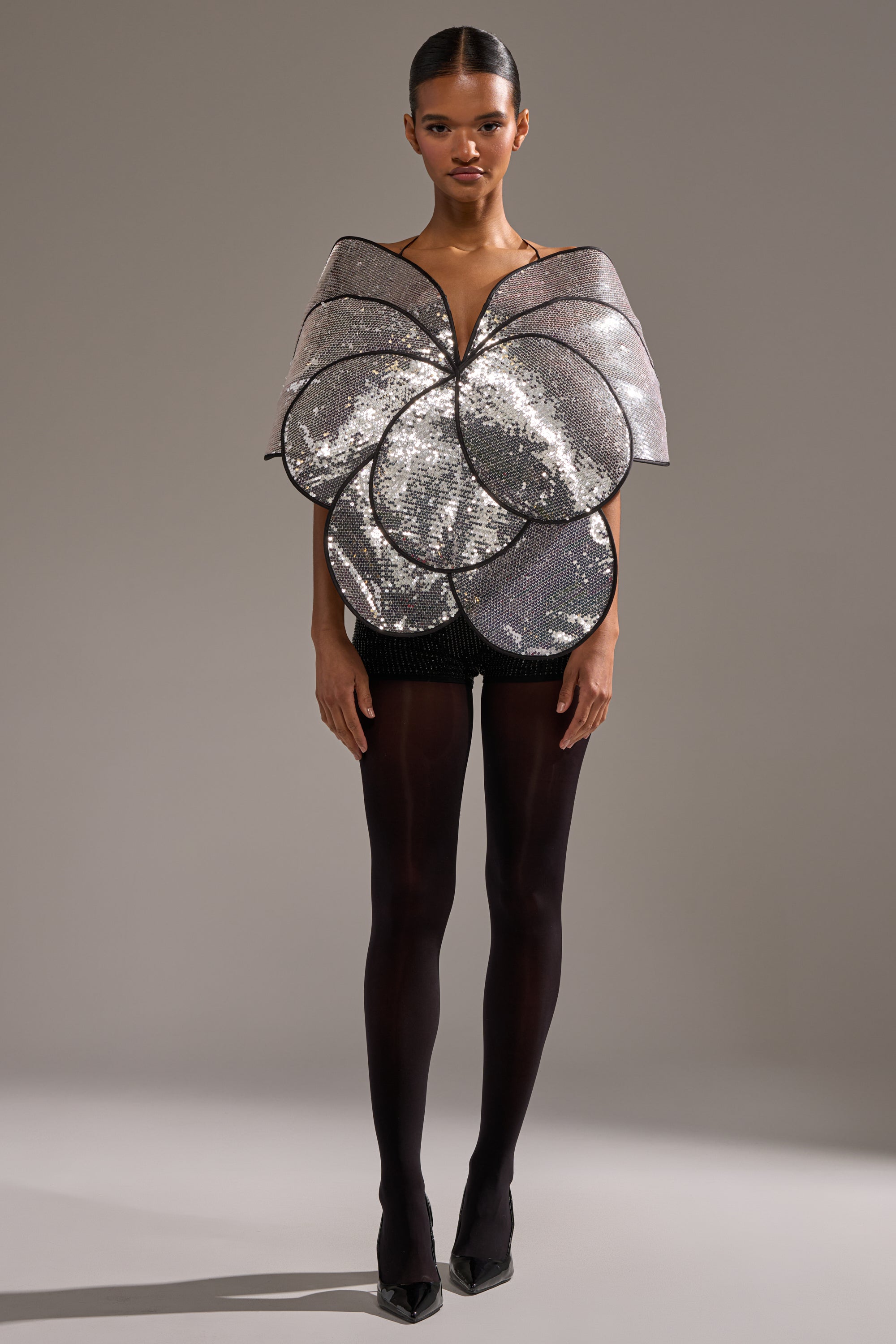 A model wears the LA DIVINA SEQUIN TOP—dramatic silver sequins shaped like oversized flower petals—paired with black shorts, sheer tights, and pointed heels against a plain light gray background. 5