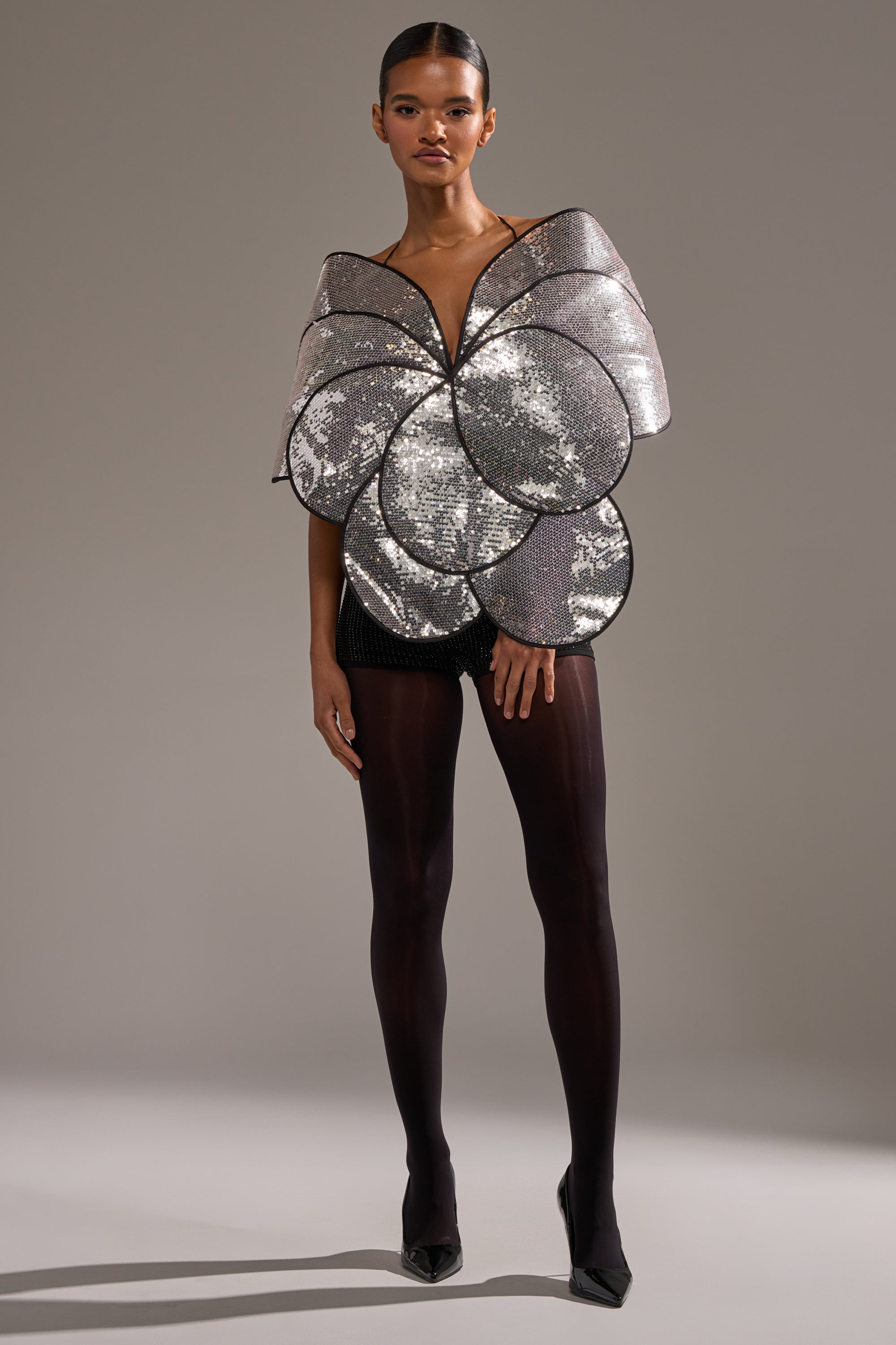 A woman wears the LA DIVINA SEQUIN TOP—dramatic silver with petal-shaped sequins—styled with black shorts, sheer tights, and pointed heels against a neutral background. 6