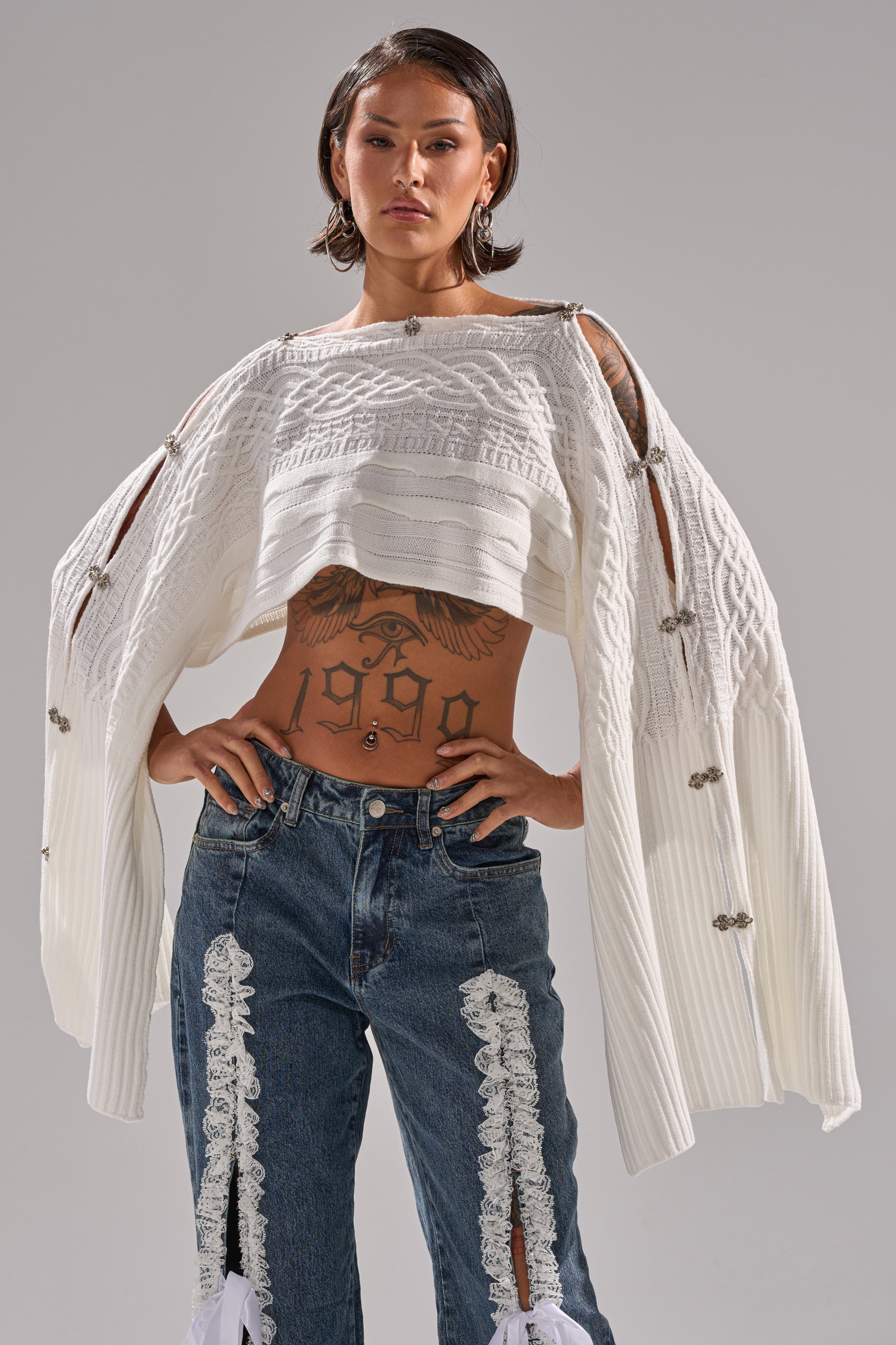 A woman confidently wears the JOAN CABLE KNIT TOP, a textured white cropped sweater with draped sleeves and metal clasps, paired with blue jeans adorned with decorative white lace panels. Tattoos are visible on her midriff and chest.