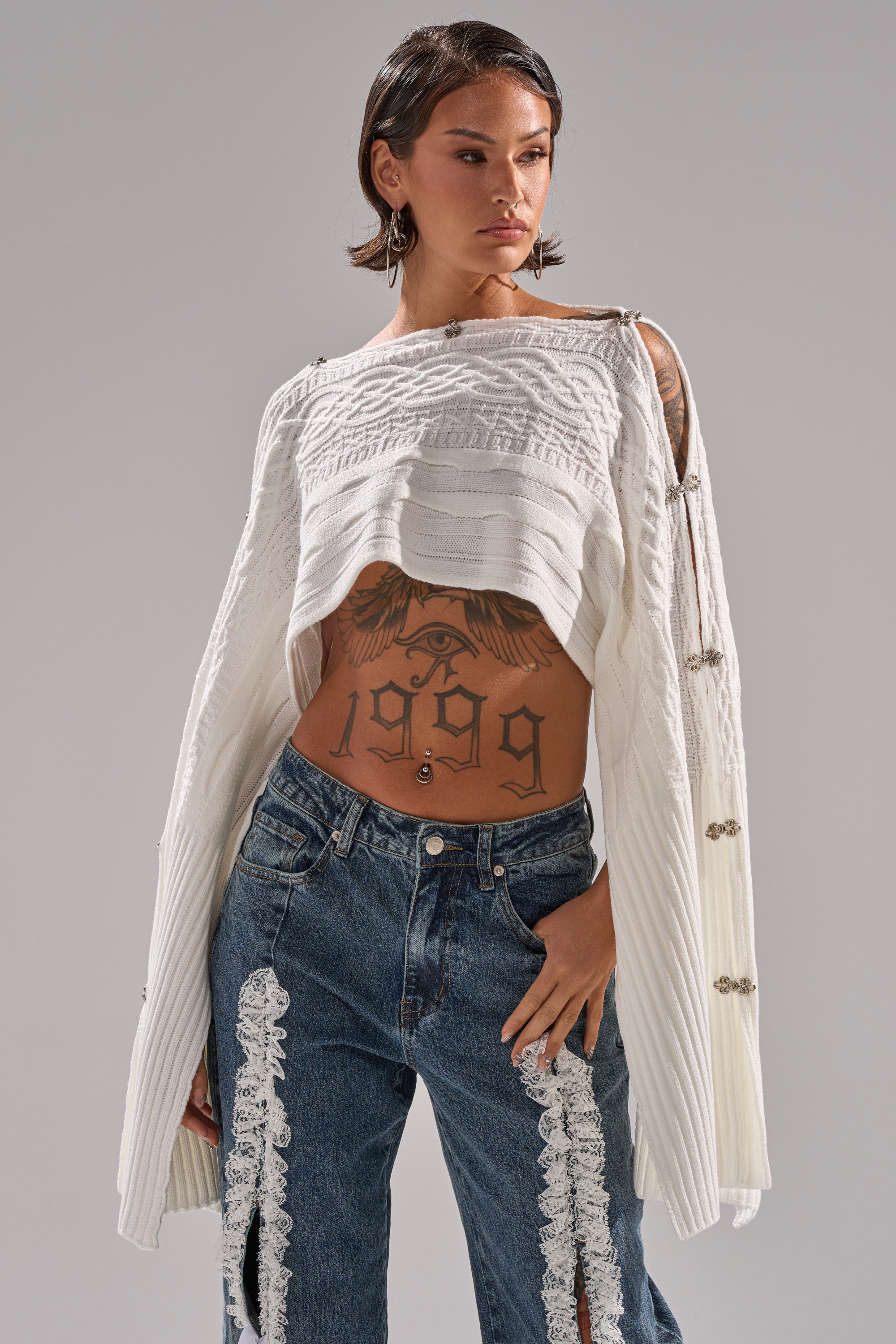 A person with short dark hair wears the JOAN CABLE KNIT TOP, an open sweater with draped sleeves, and blue jeans with lace accents. Tattoos on their stomach and arms include a large eagle and "1999" on their torso.