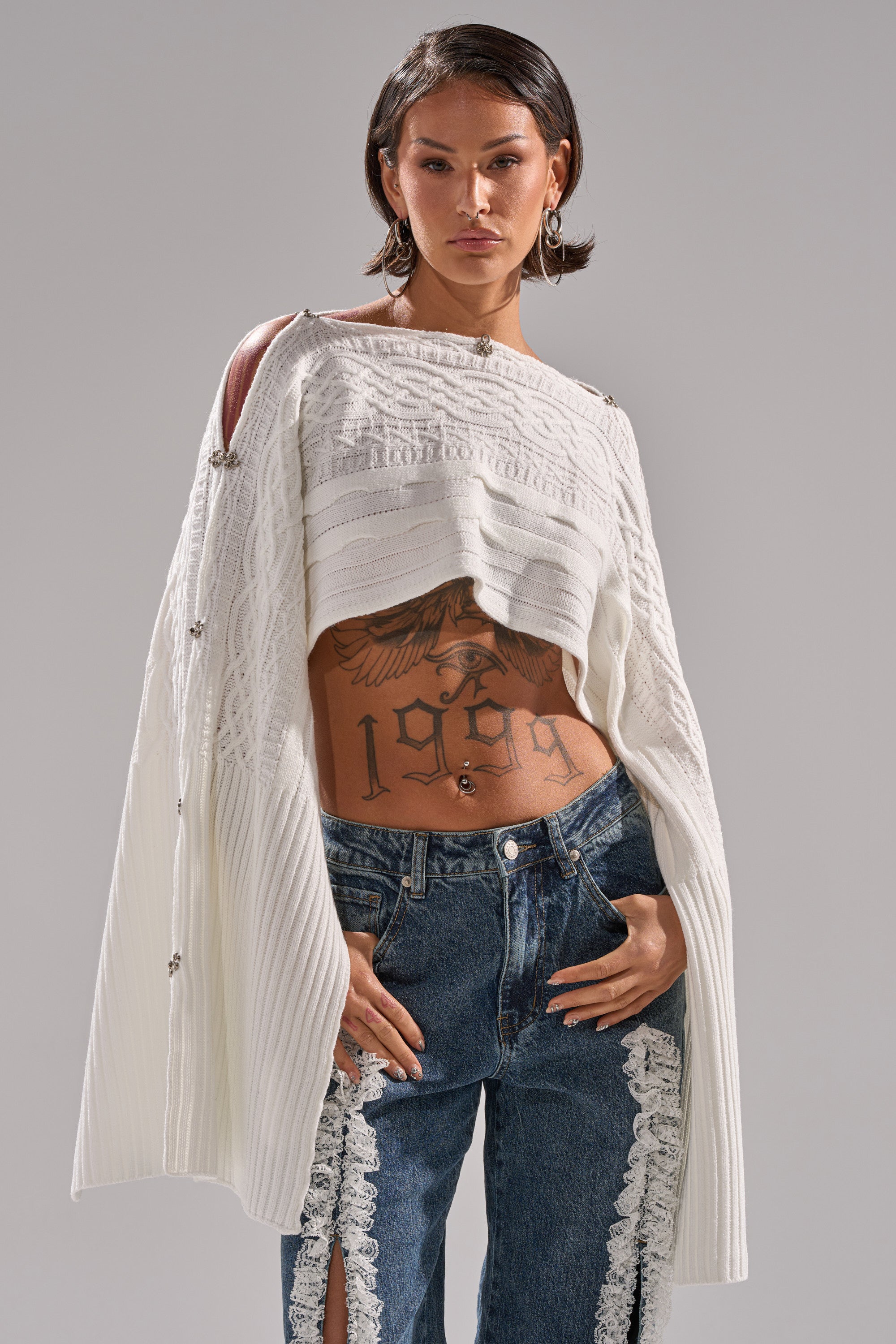 A person with short dark hair wears the JOAN CABLE KNIT TOP by AKIRA Label, a white asymmetrical cable knit style, paired with blue jeans with white lace trim. Their visible midriff reveals tattoos, including a large eagle and "1959.