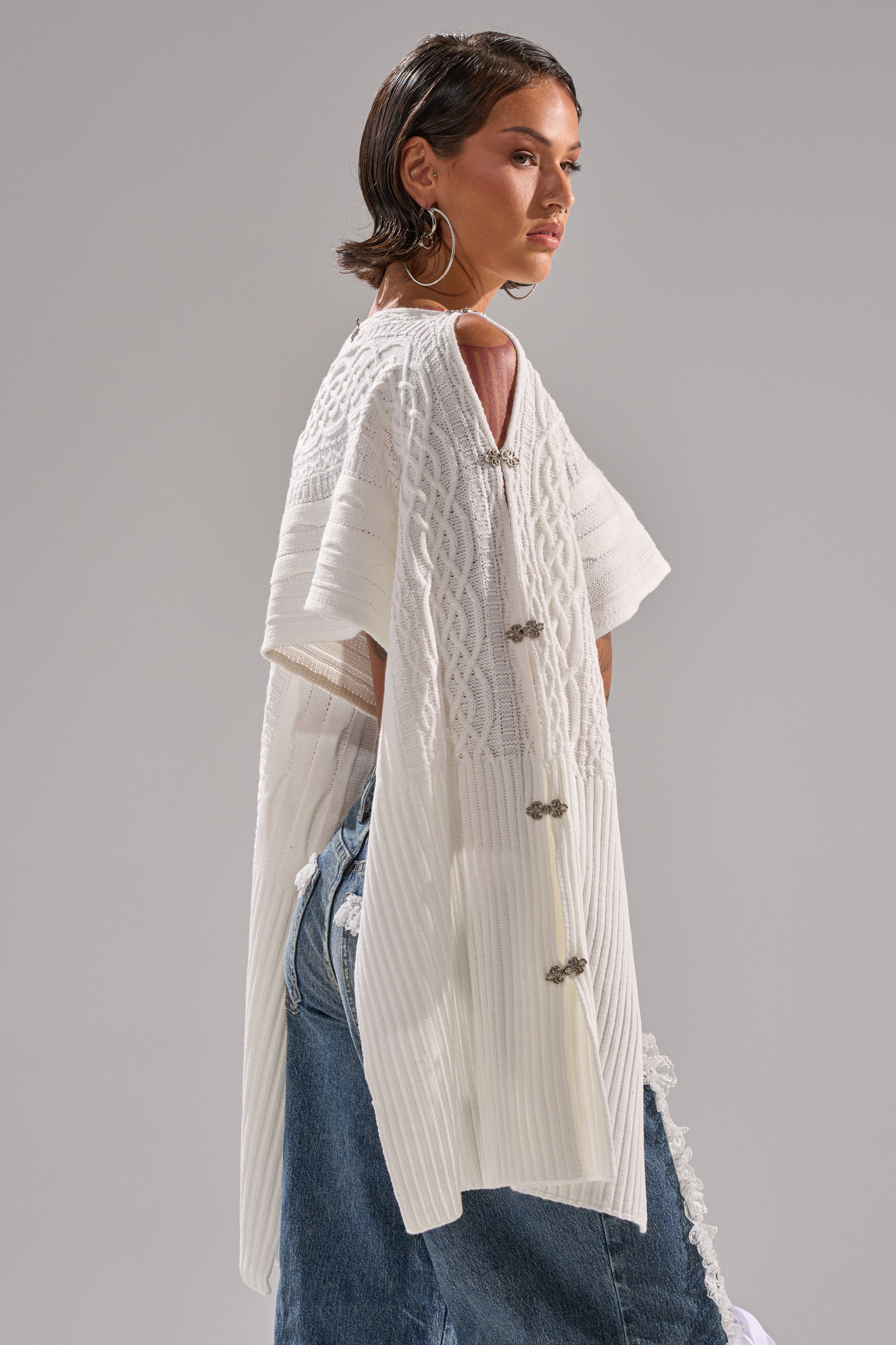 A woman with short, slicked-back hair and large hoop earrings models the JOAN CABLE KNIT TOP—featuring a loose fit, dramatic sleeves, and decorative back clasps—paired with light blue jeans.