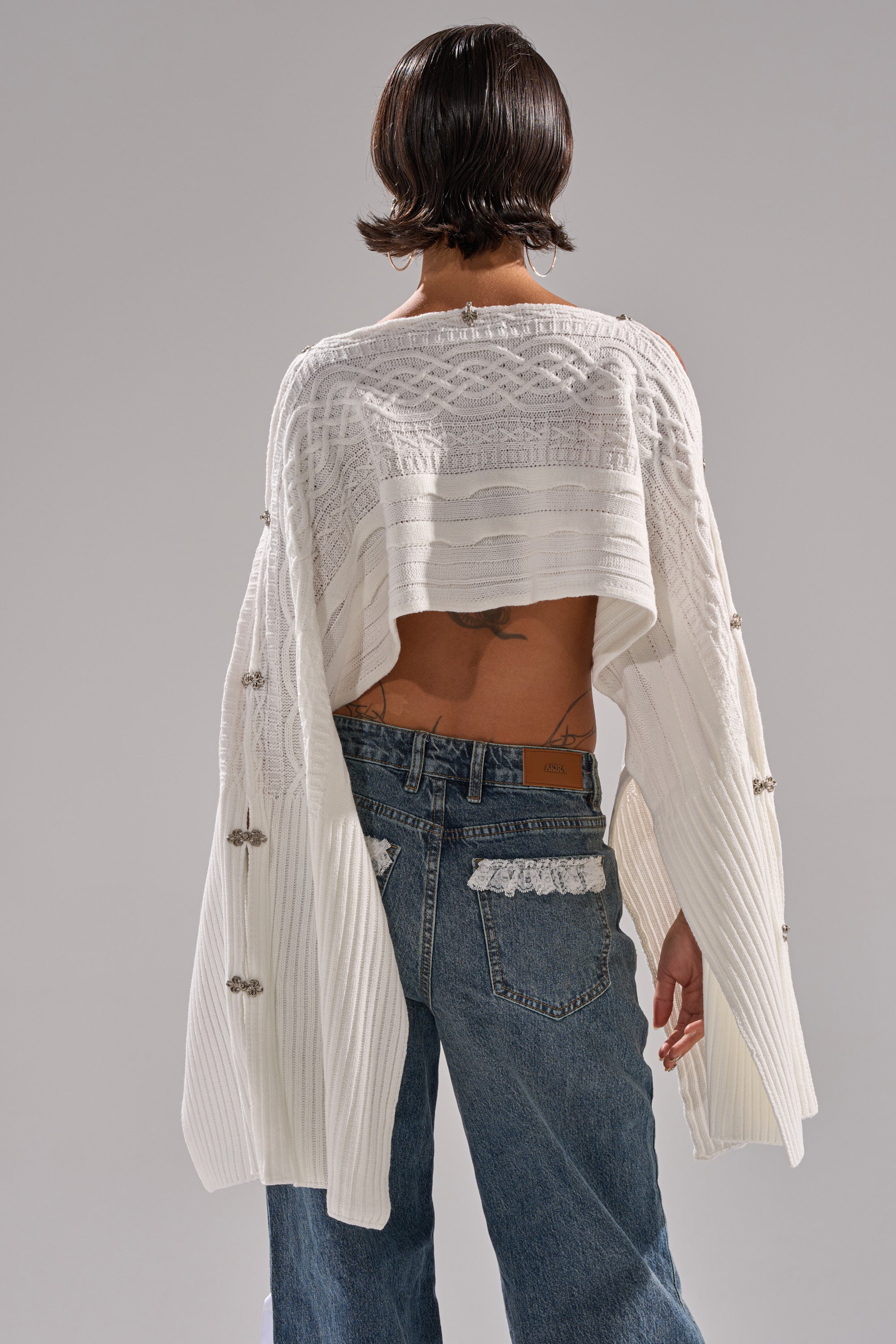 A person with short dark hair is seen from behind wearing the JOAN CABLE KNIT TOP—a white, cropped cable knit sweater with decorative buttons, dramatic sleeves, and blue jeans. A tattoo is visible on their lower back above the jeans.