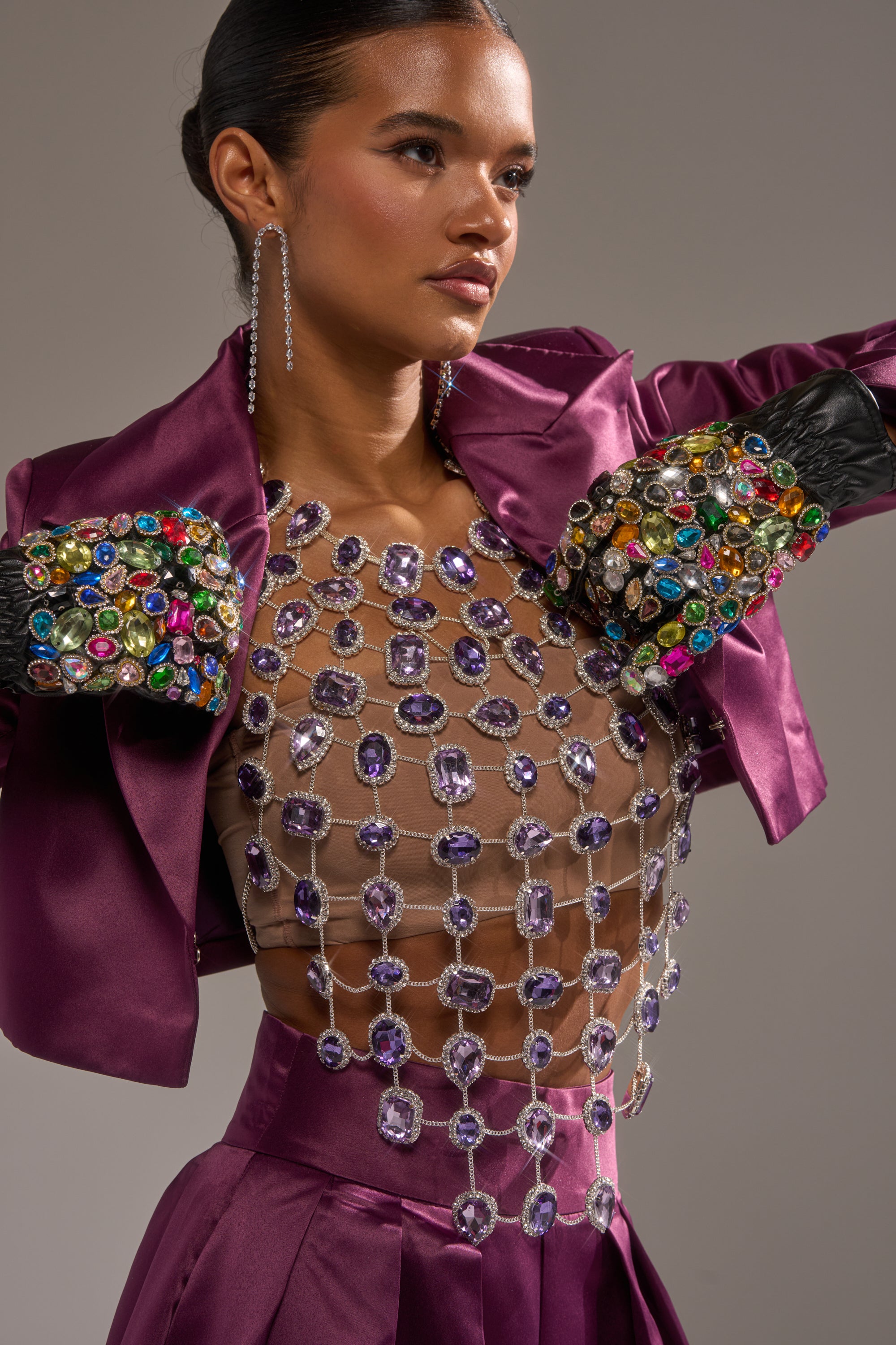A woman poses in the LOVER GIRL RHINESTONE CHAIN TOP, featuring jeweled embellishments and a grid of large crystals. She pairs it with long, sparkling earrings and wears her hair slicked back. 0
