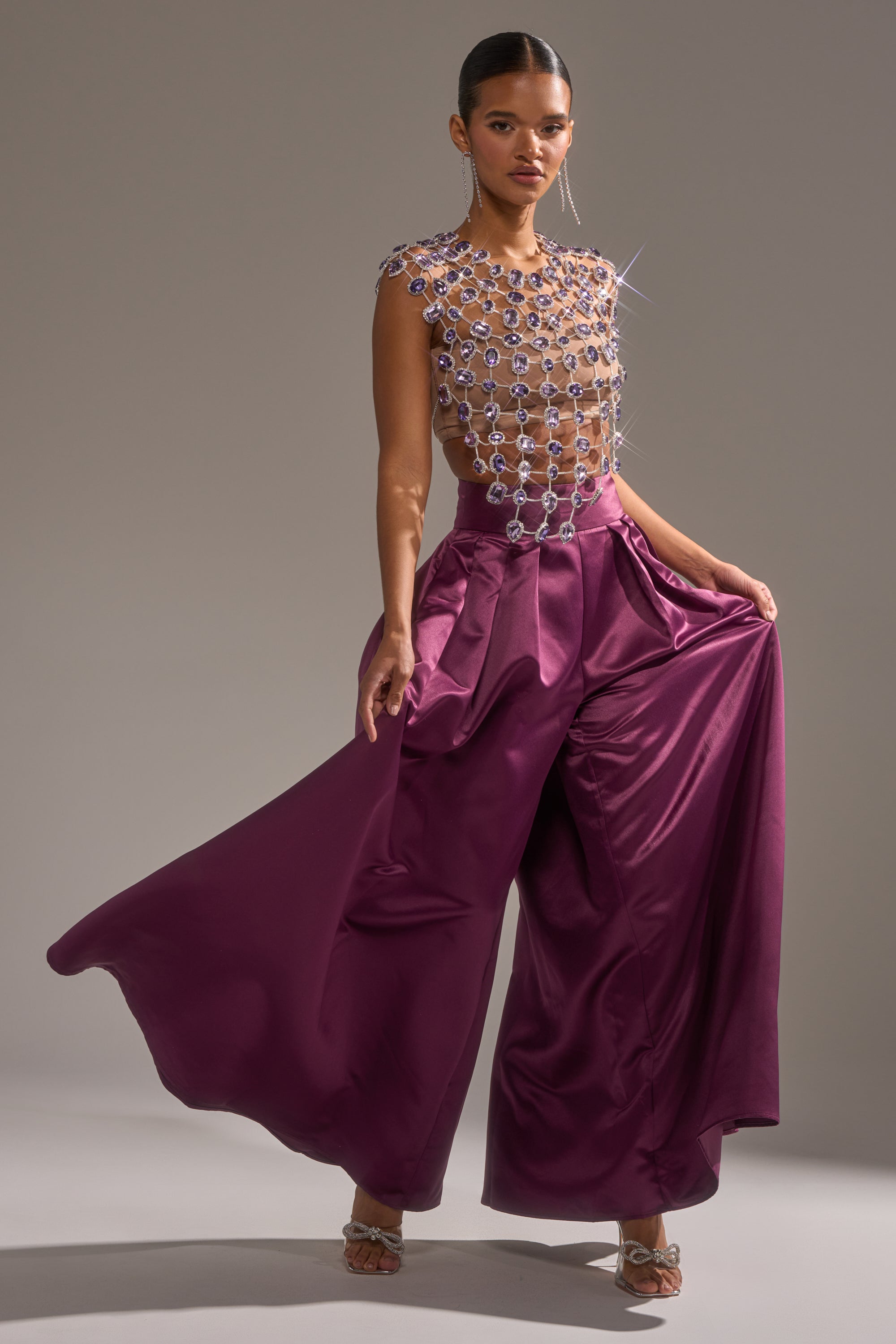 A woman poses confidently in the LOVER GIRL RHINESTONE CHAIN TOP and wide-legged, high-waisted magenta satin pants, striking a statement silhouette as she holds out one side of her flowing pants. 9