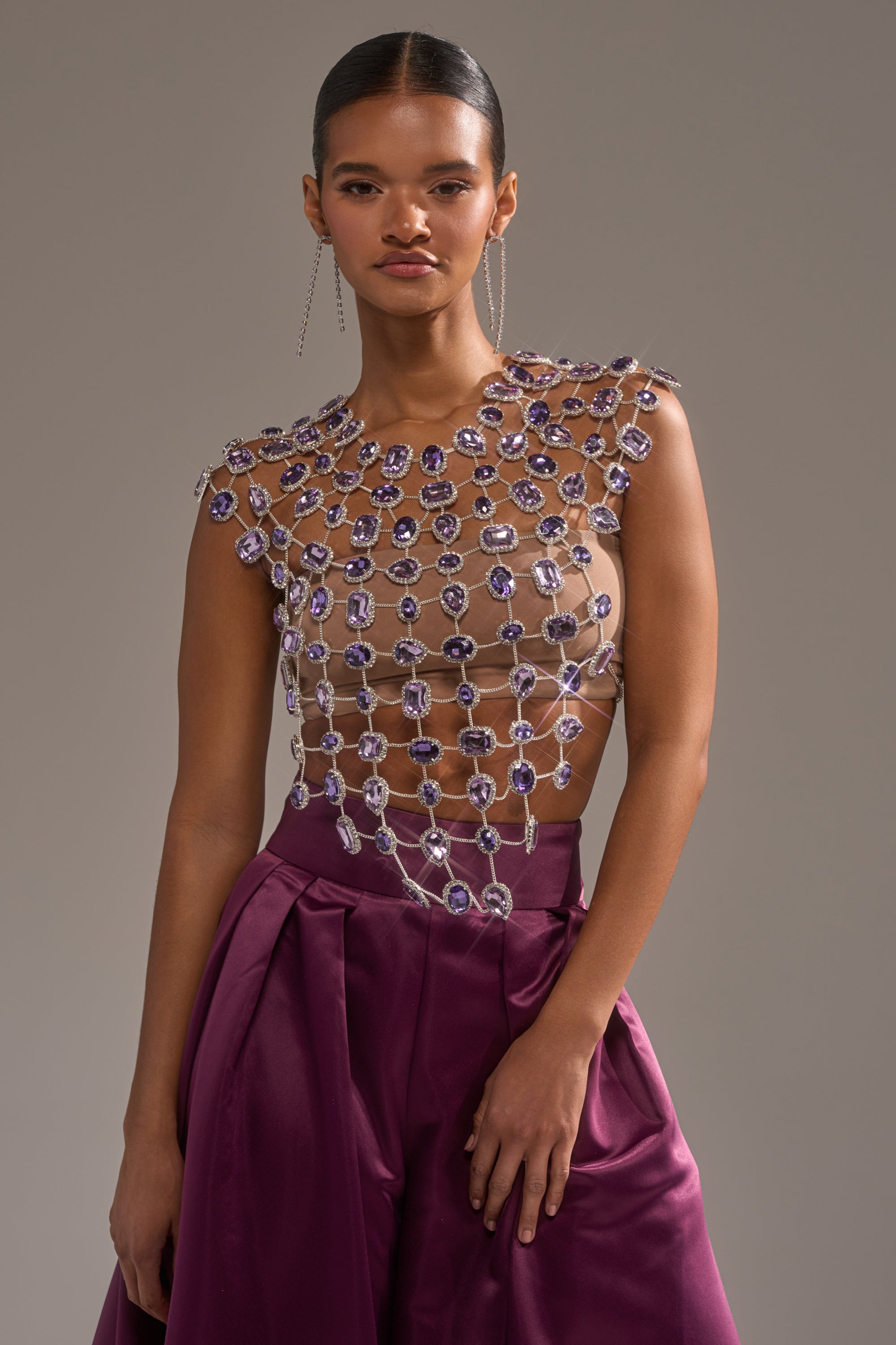 A woman wears the LOVER GIRL RHINESTONE CHAIN TOP over a nude base, paired with high-waisted deep purple satin pants. Her hair is slicked back and she accessorizes with long, dangling earrings. 1