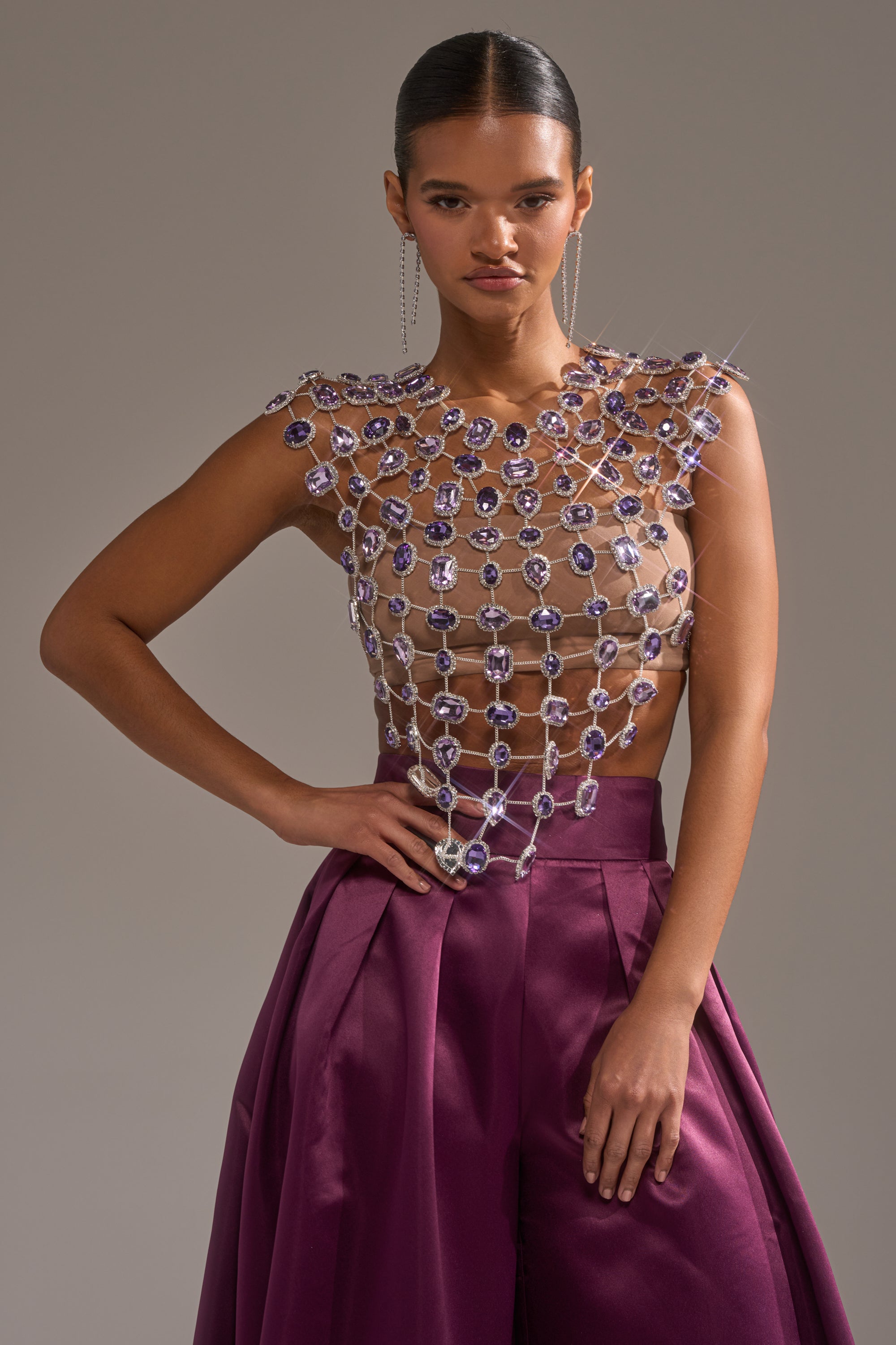 A woman poses confidently in the LOVER GIRL RHINESTONE CHAIN TOP, featuring sparkling crystals, paired with high-waisted deep purple satin pants. Her slicked-back hair and long, delicate earrings complete the look against a neutral background. 3
